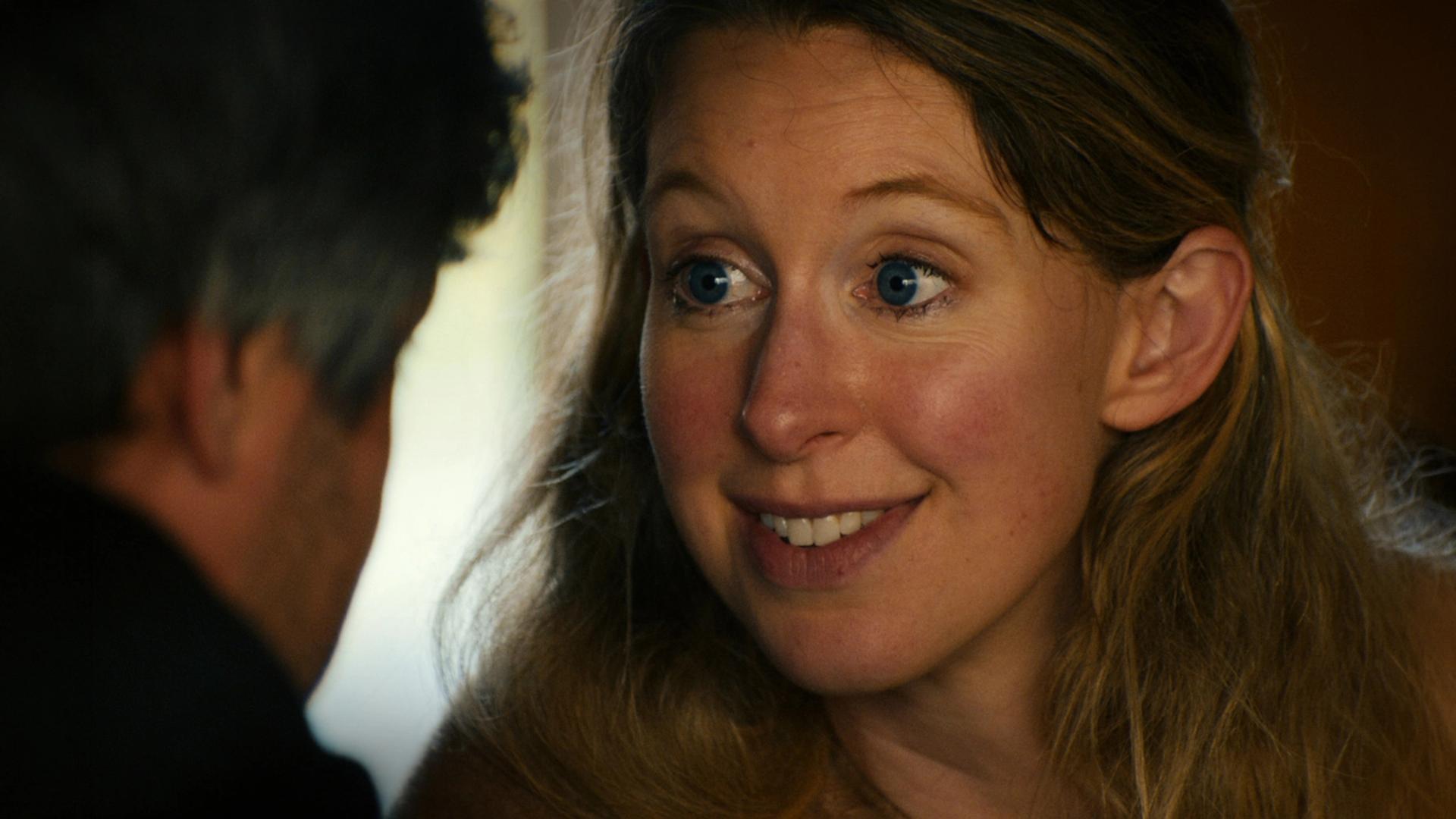 a wide-eyed, smiling woman with long reddish-brown hair looking at an out-of-focus person facing her in the foreground, warmly lit indoors