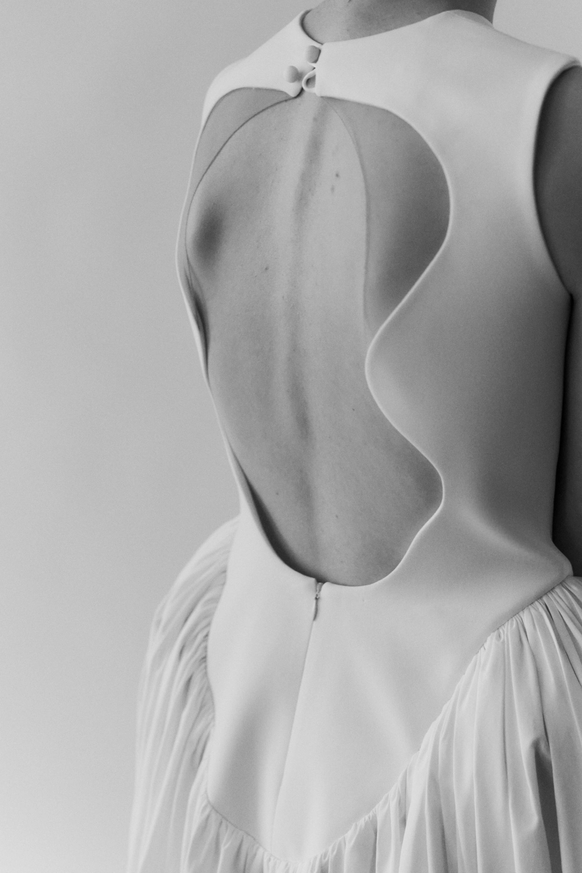 A black and white studio photograph of the back of a woman wearing a white dress with a ruffled drop waist and curved open back detail