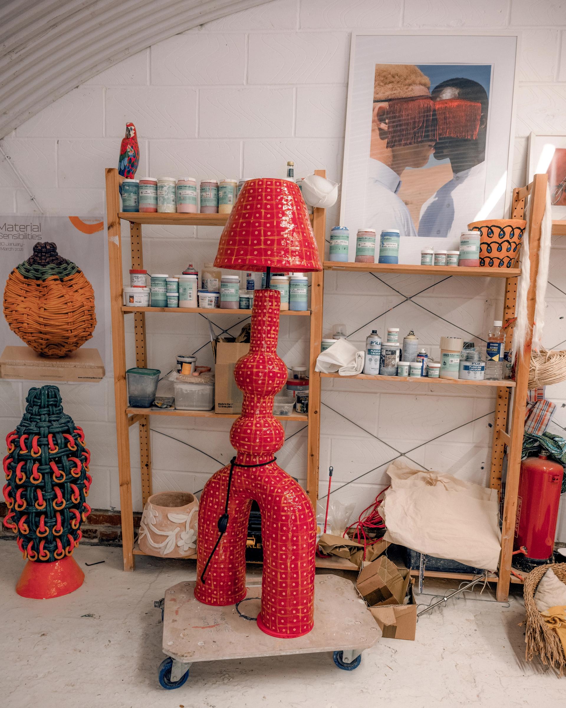 An interior space with a bold red patterned floor lamp on a trolley, wooden shelves of paint tins, and colorful woven basket sculptures on the floor.