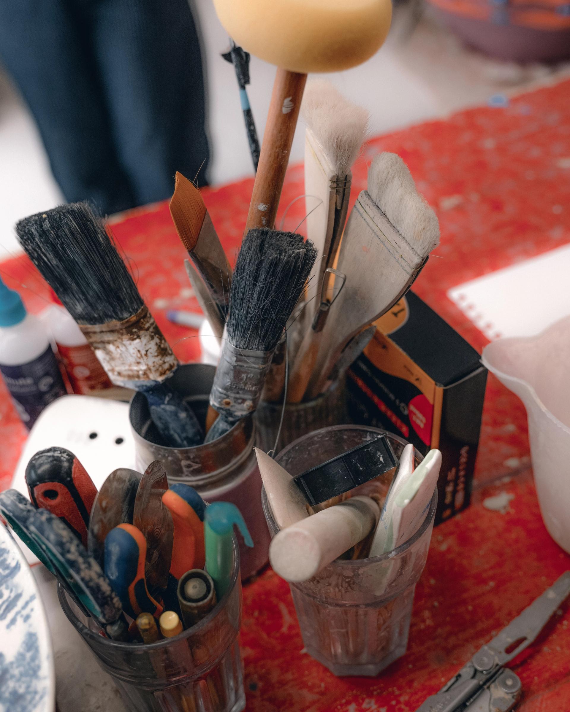 Glasses with tools including paintbrushes and writing implements sit on top of a worn red work surface.