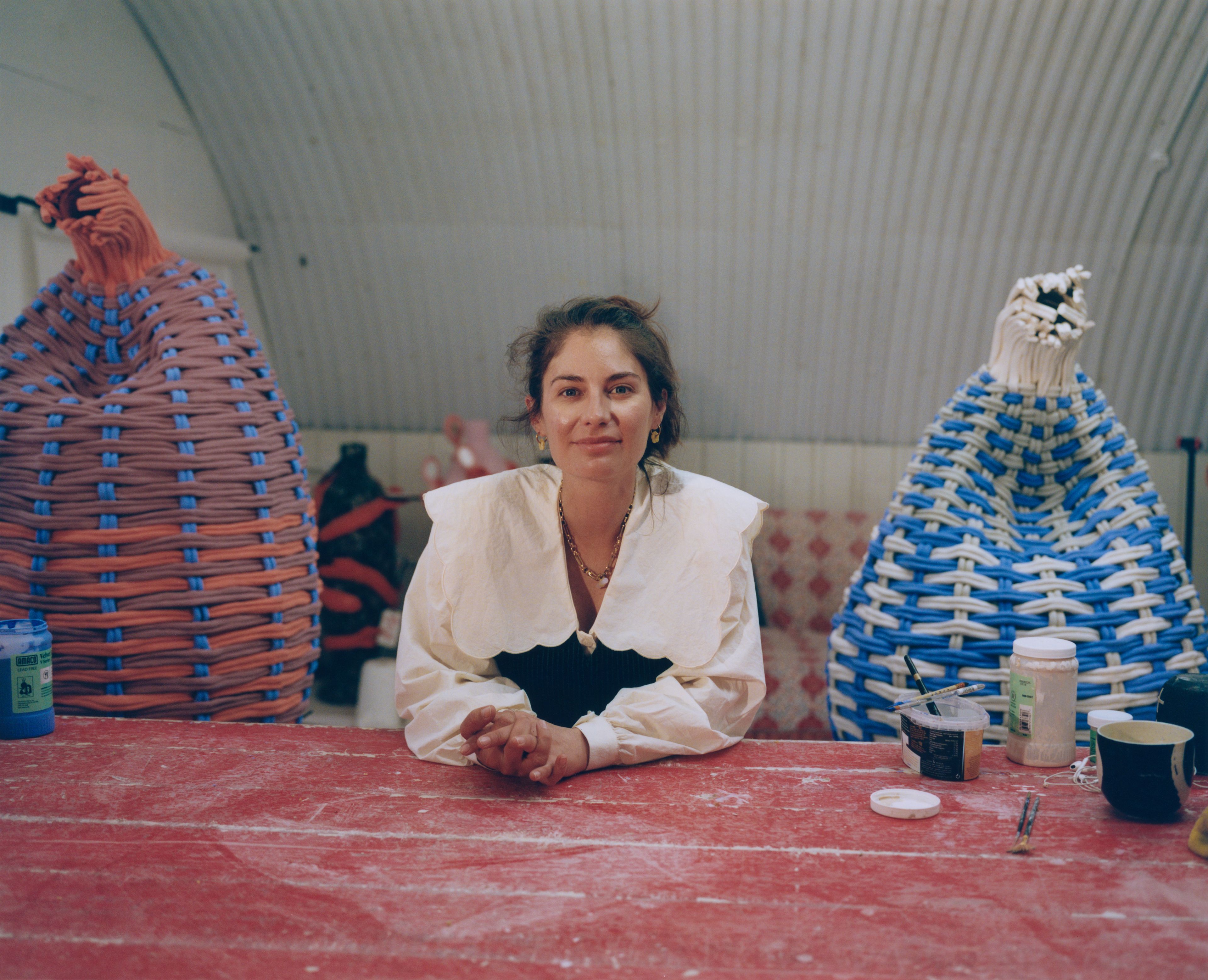 A woman in a white blouse with a wide collar rests her arms on a worn red surface, with large woven basket sculptures visible behind her.