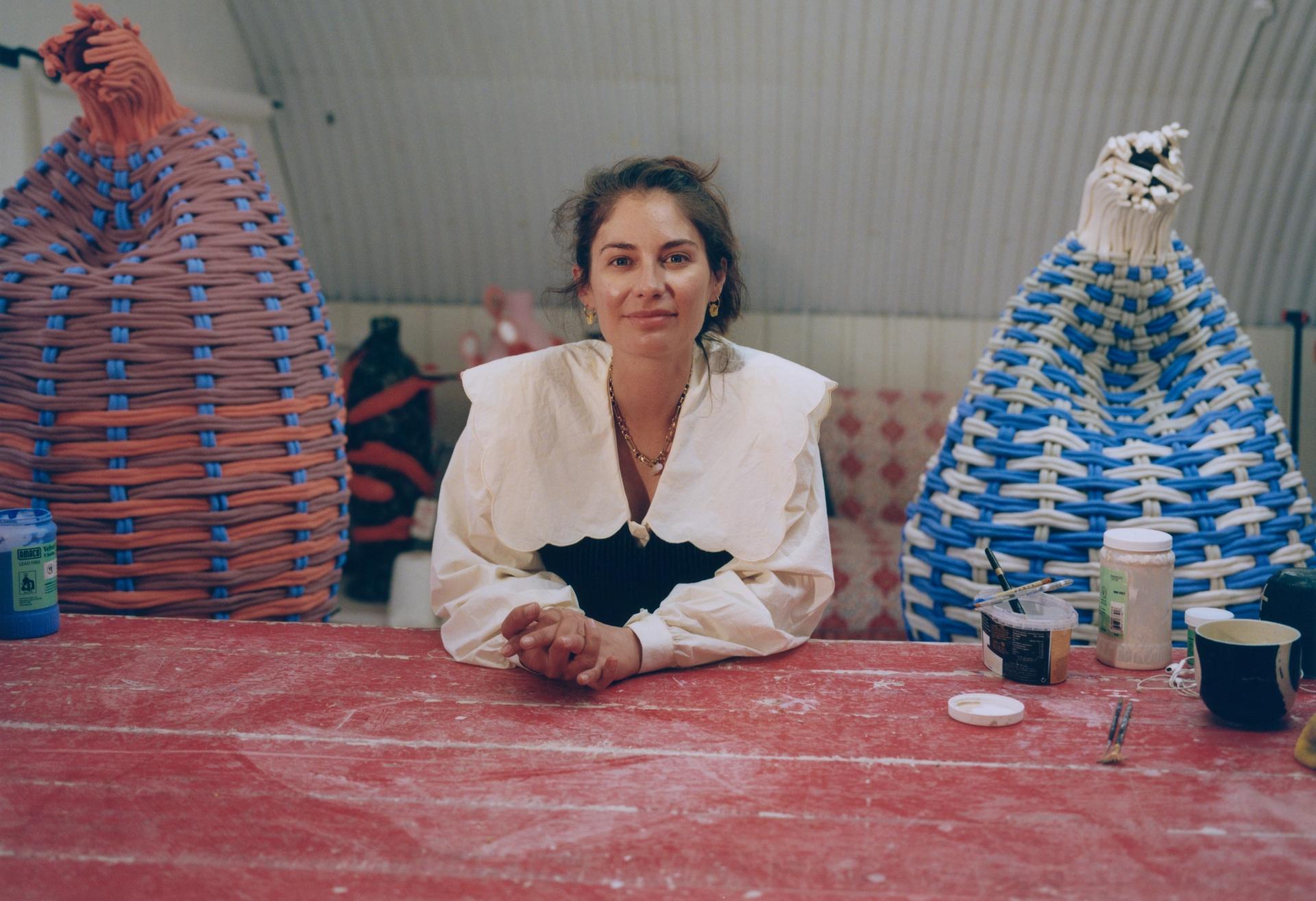 A woman in a white blouse with a wide collar rests her arms on a worn red surface, with large woven basket sculptures visible behind her.