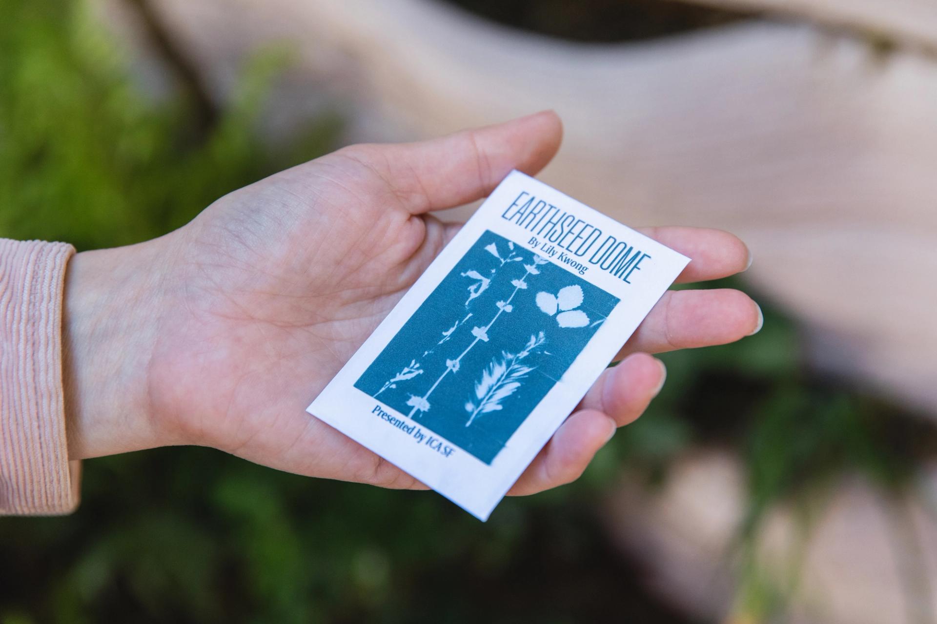 A close-up shot of a hand holding a white paper packet printed with a blue cyanotype and reading "Earthseed Dome by Lily Kwong Presented by ICA SF"