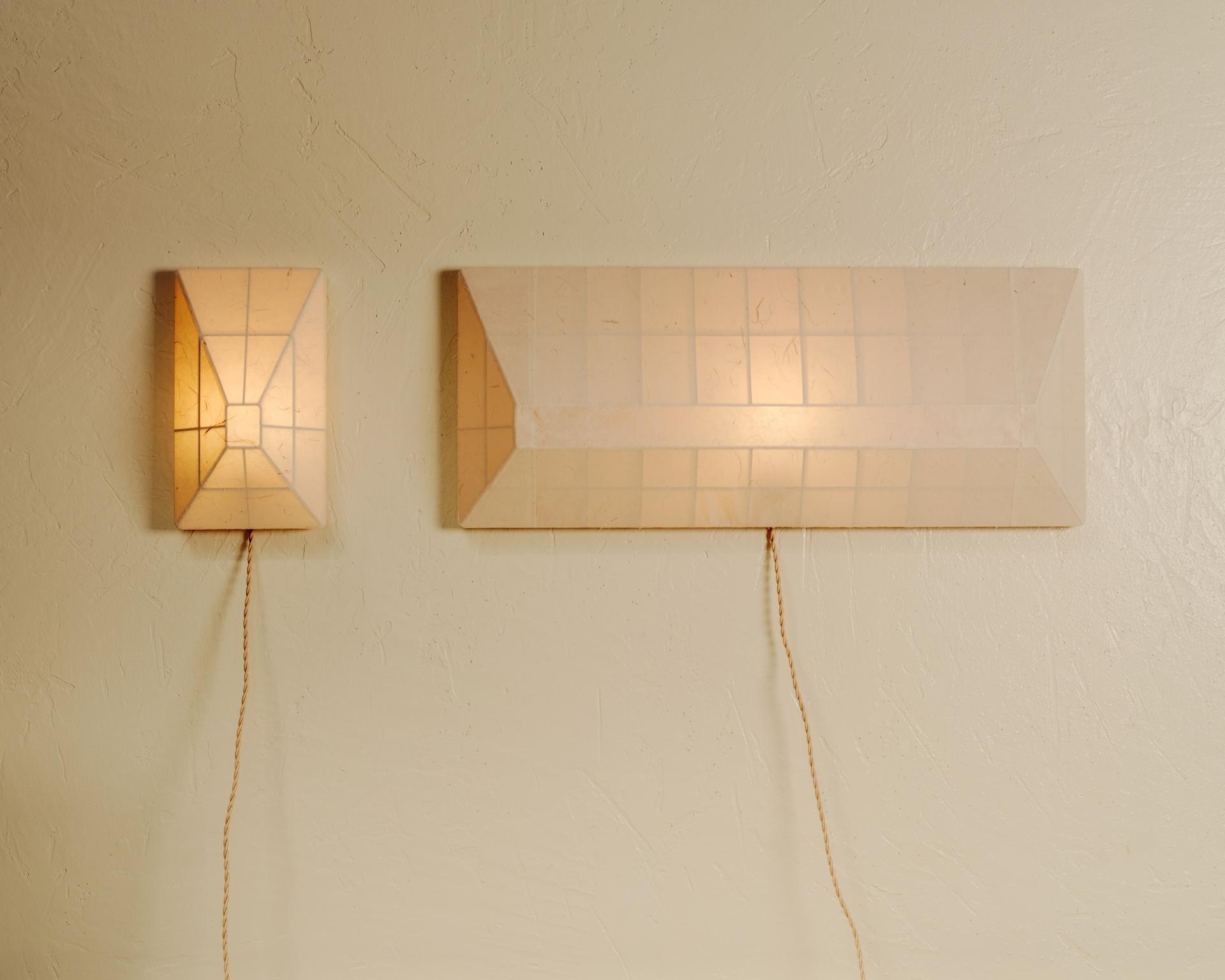 Two geometric wall sconces with warm amber light hang above a cream upholstered bench against a textured off-white wall.