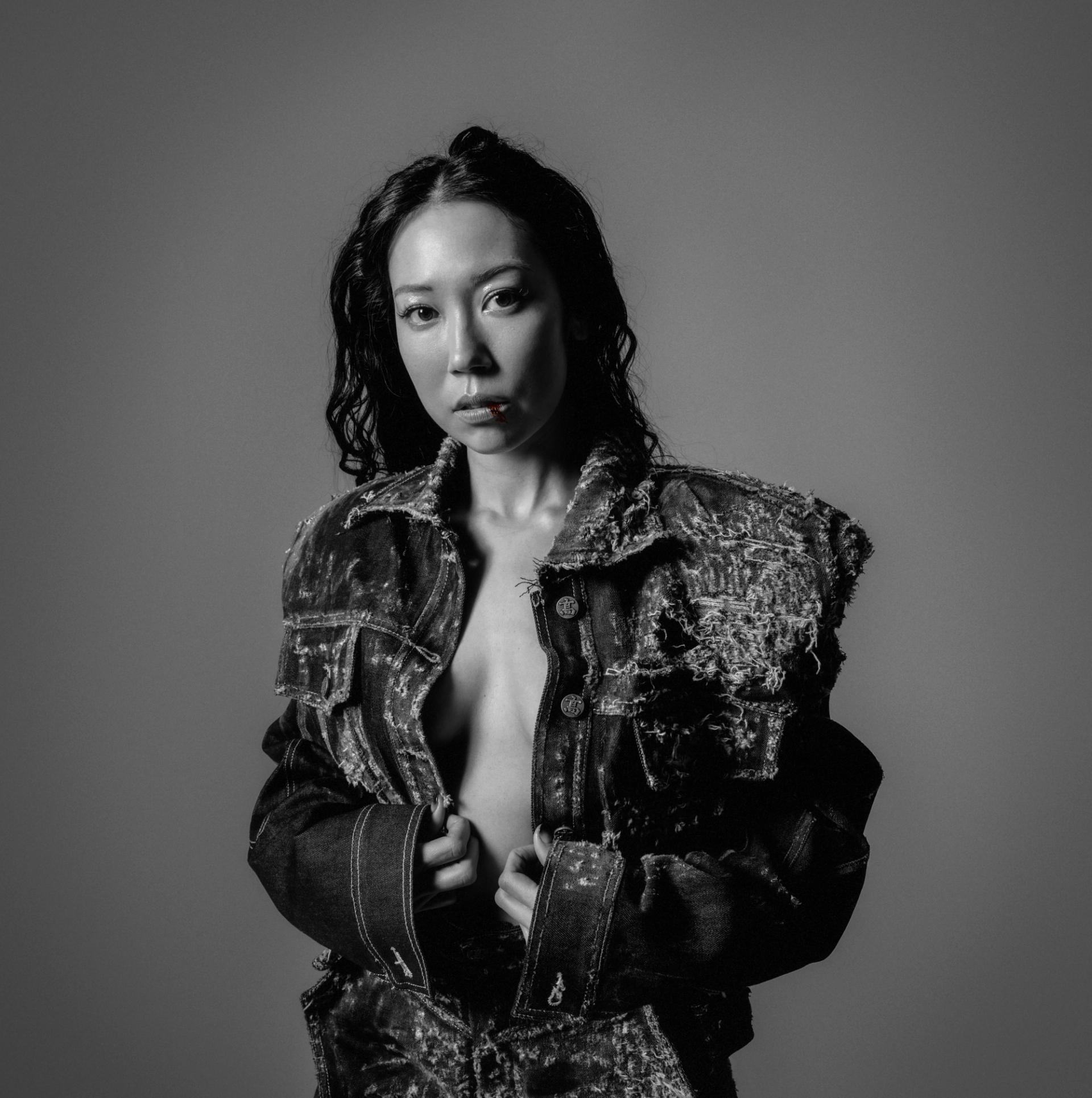 A black and white studio portrait of a woman in a heavily distressed denim jacket, gazing directly at the camera against a gray background
