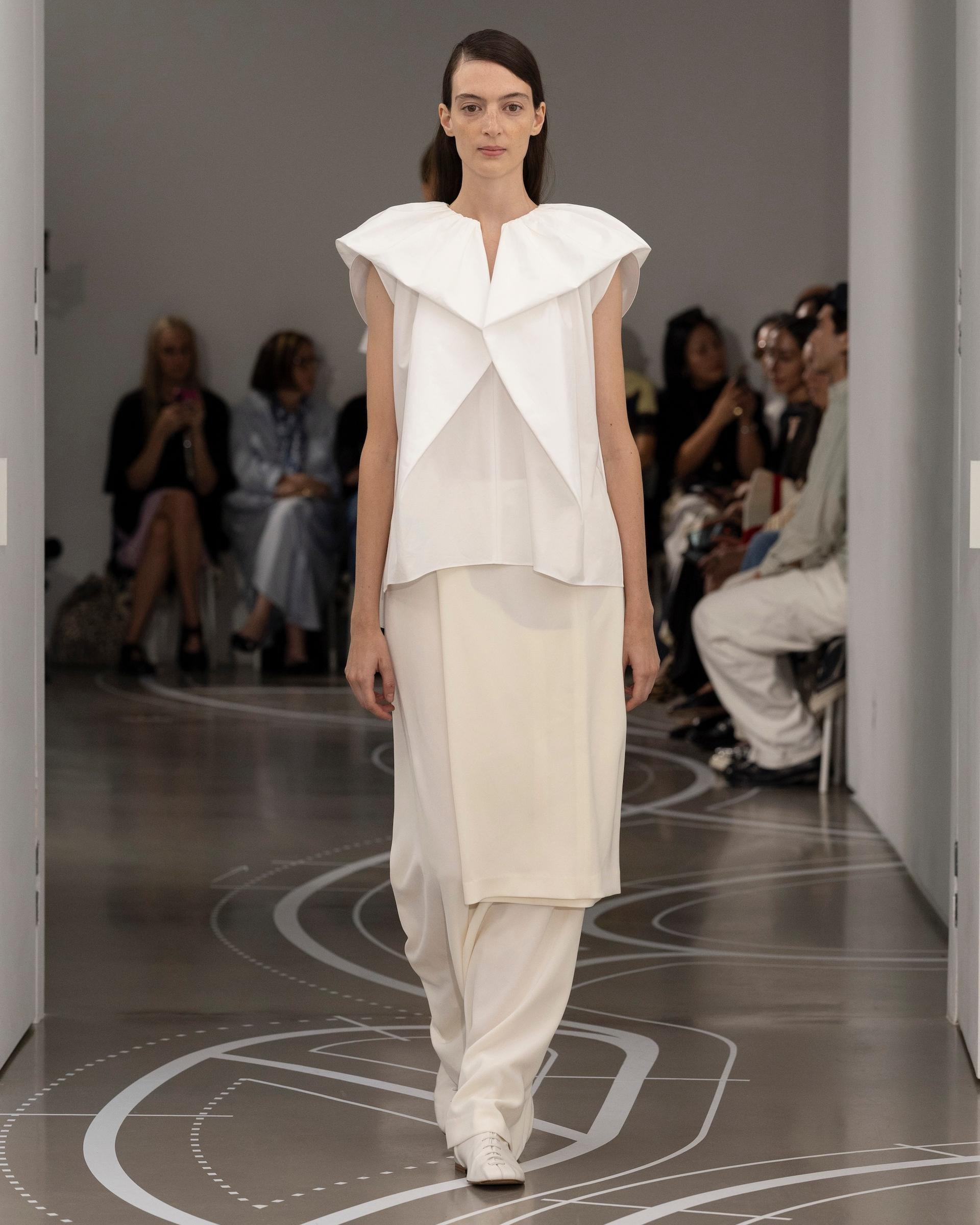 A model walks a runway in an all-white look featuring a structured origami-fold top and wide-leg trousers, with seated guests visible on either side