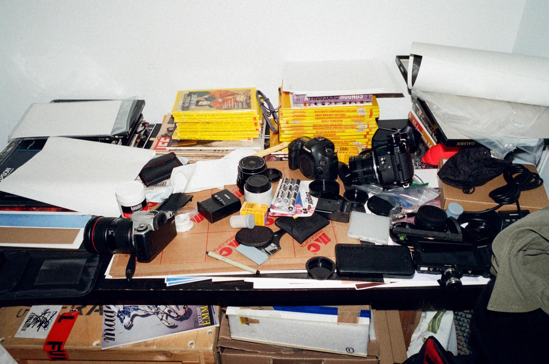 A messy desktop covered with cameras, lenses, and miscellaneous magazines and office supplies.