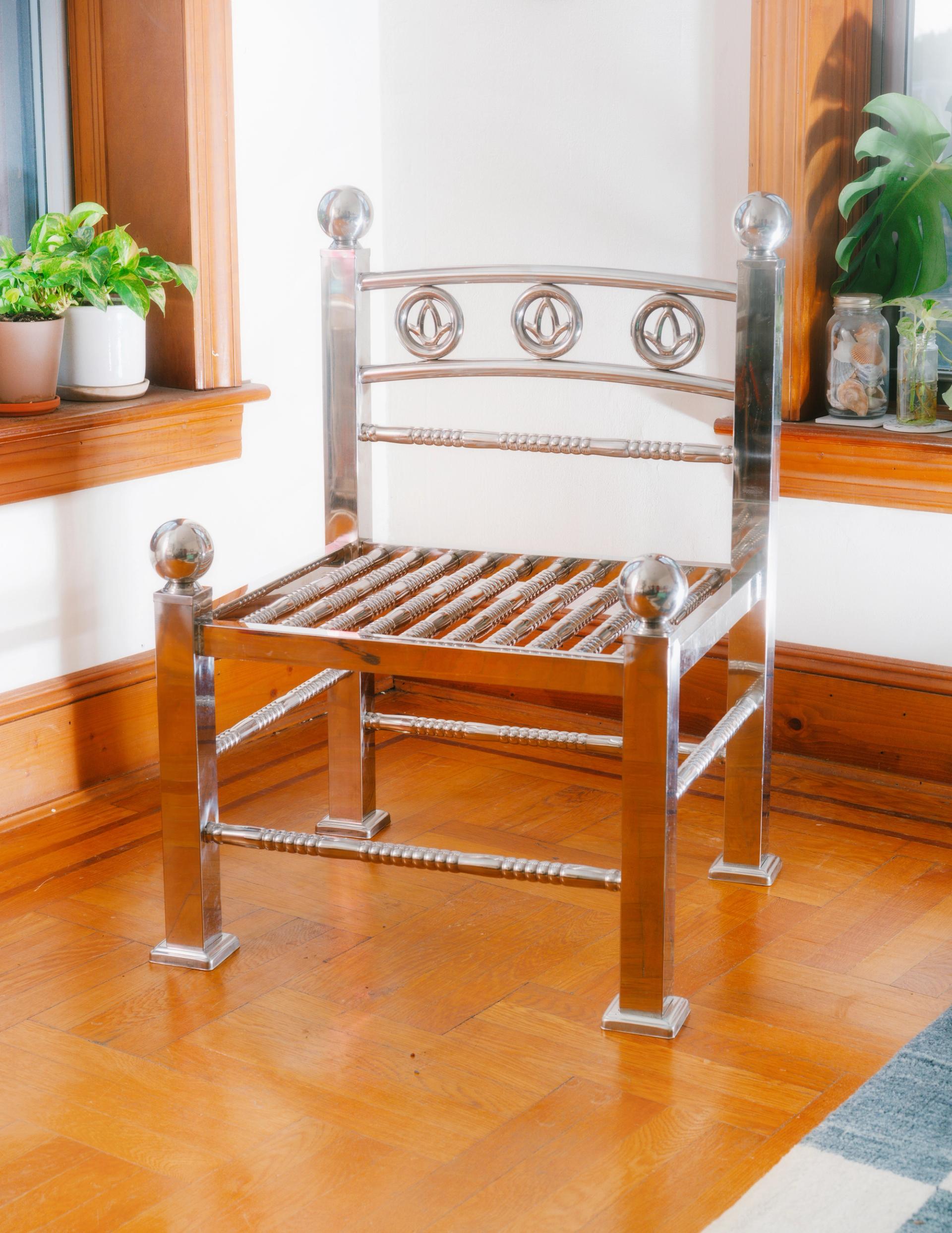 A shiny stainless steel chair with ornate design elements sits in the corner of a sunlit room.
