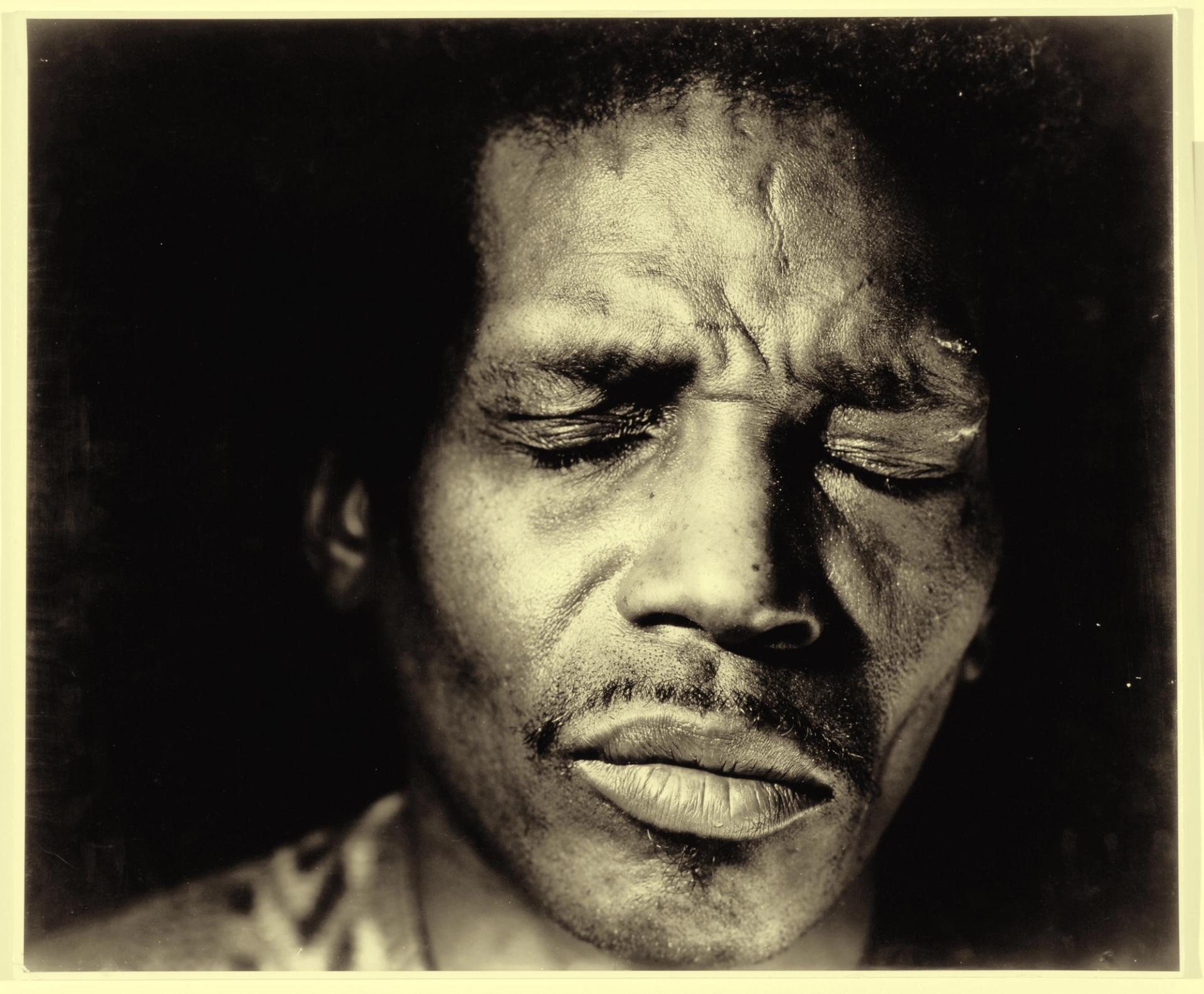 Close-up black-and-white portrait of man with eyes closed, face dramatically lit against dark background.