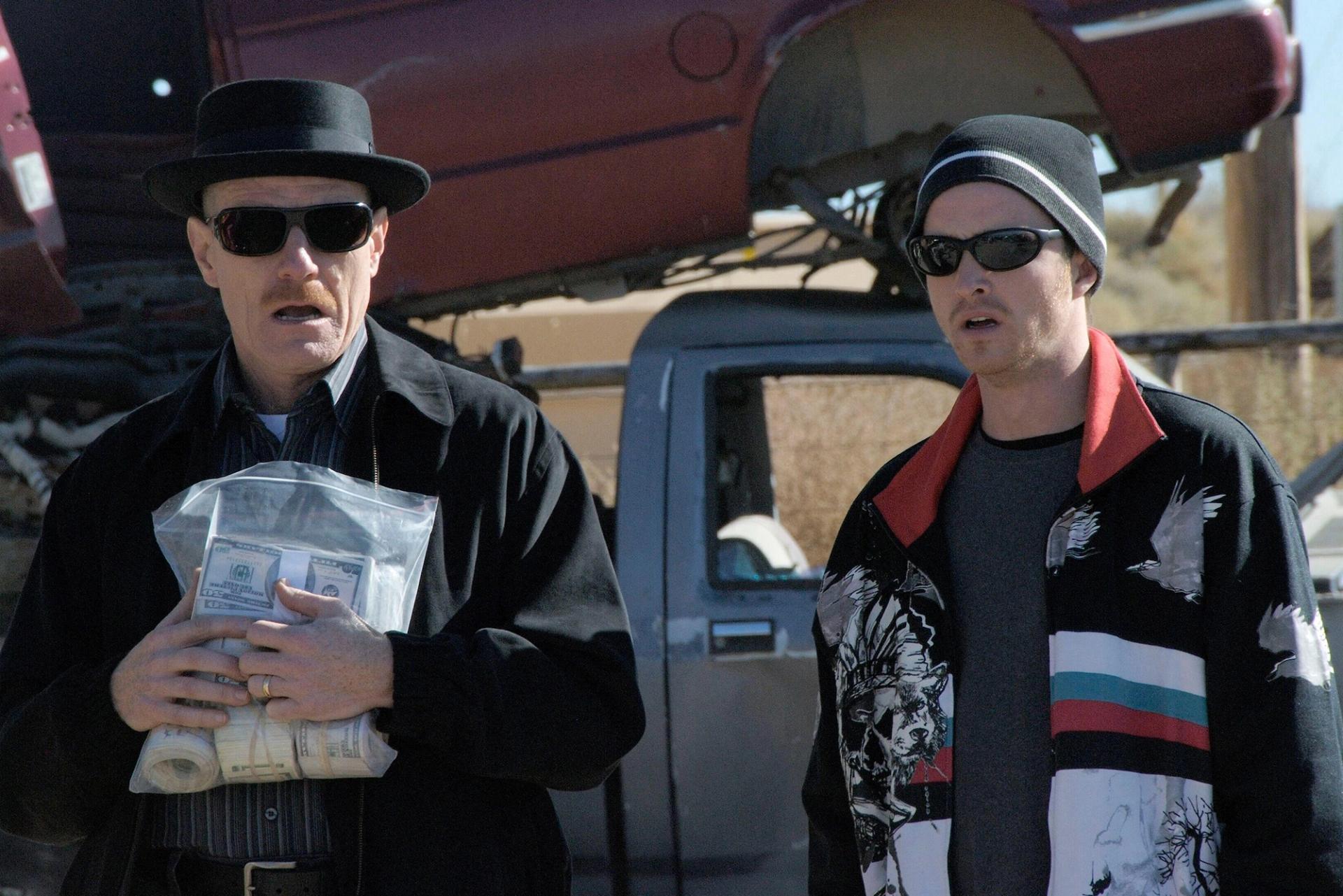 Two men in sunglasses standing outdoors near parked vehicles, one in a black coat and hat holding a bag of cash, the other in a striped jacket and beanie