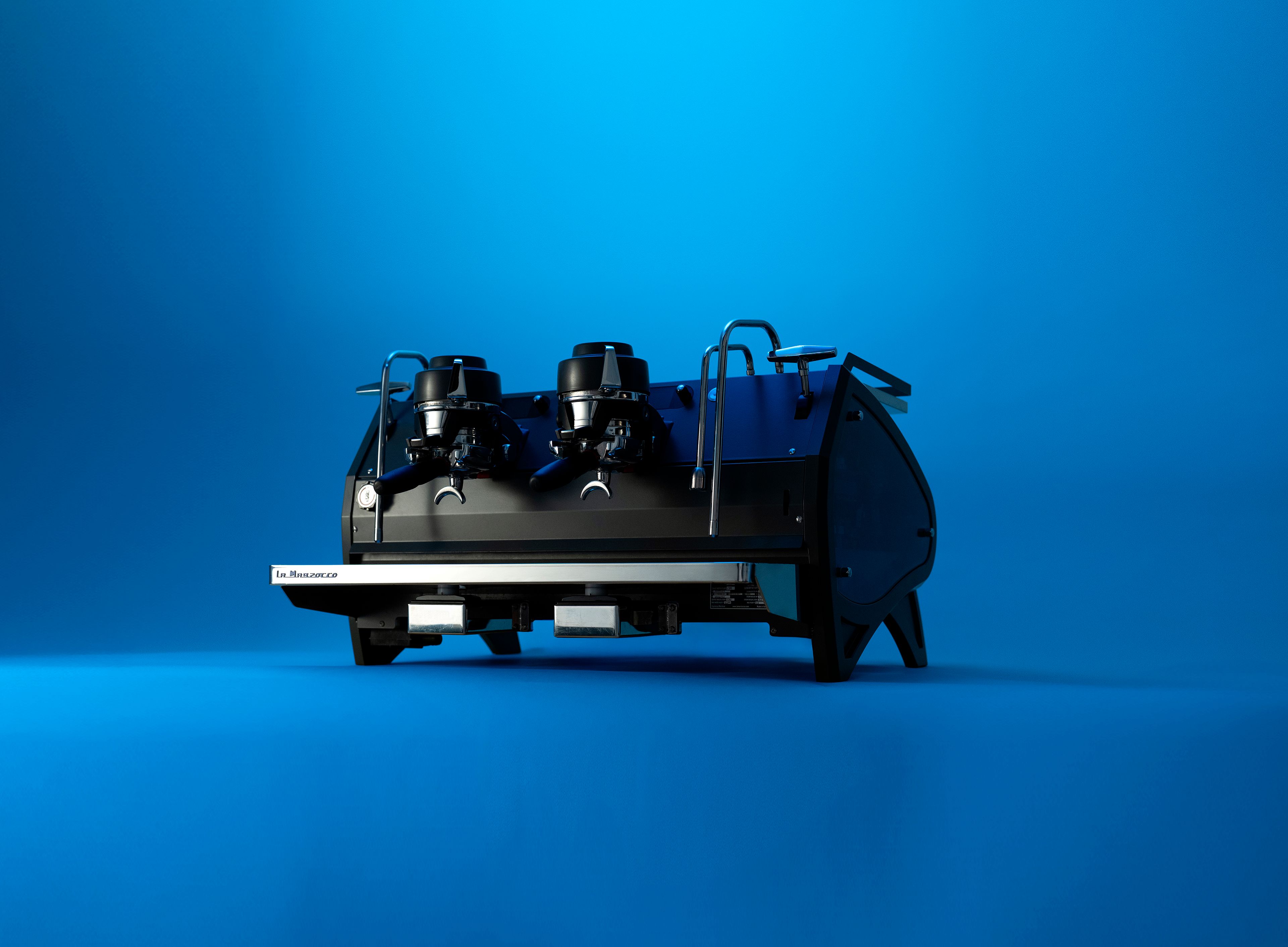 A La Marzocco two-group espresso machine in matte black, photographed against a vivid blue studio background.