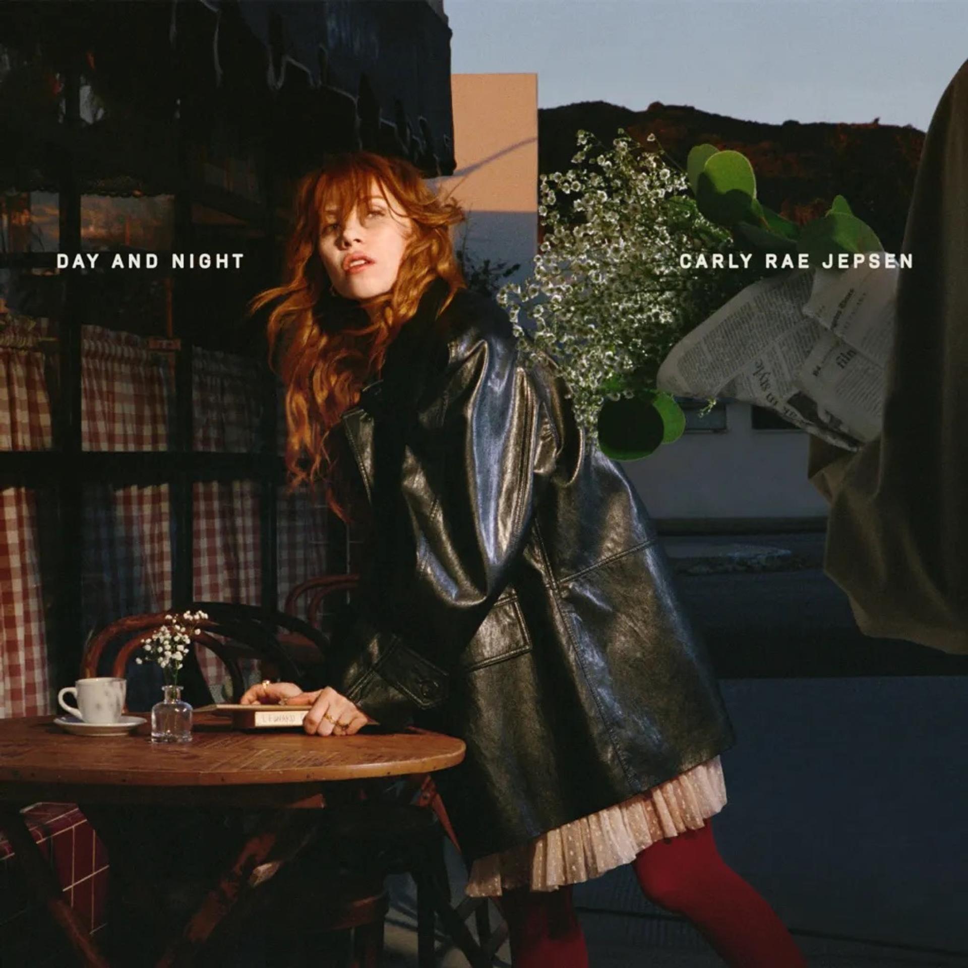 Album cover for "Day and Night" by Carly Rae Jepsen: a woman with long red hair in a black leather jacket and red tights leans against an outdoor café table, with white flowers and a hand holding a newspaper visible behind her.
