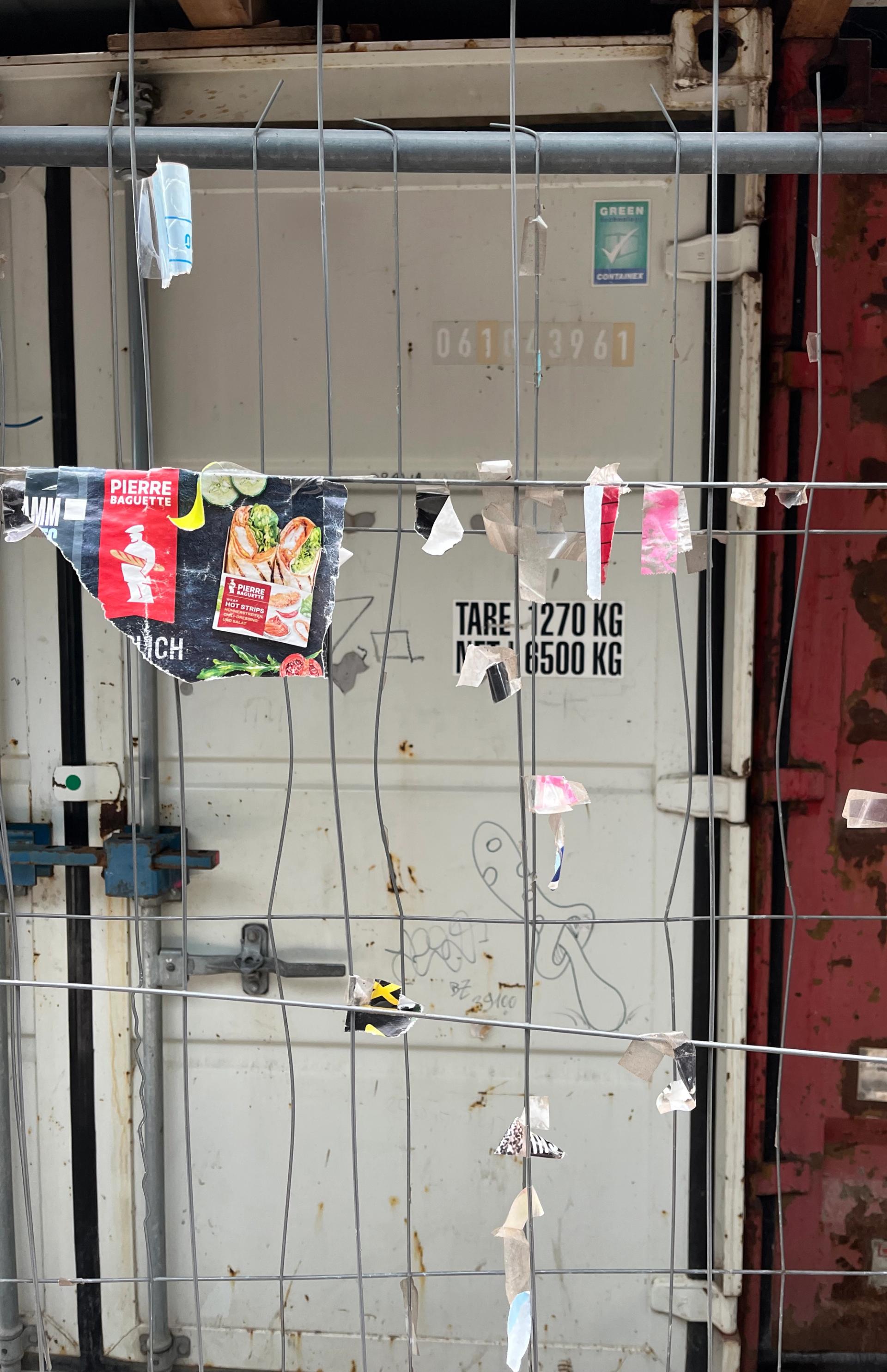 Photo of scraps of paper taped to a gridded wire barrier, in front of a shipping container.