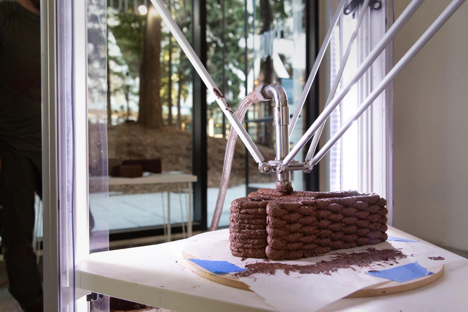 A robotic arm extrudes layers of dark clay or earth material into a small block form on a worktable