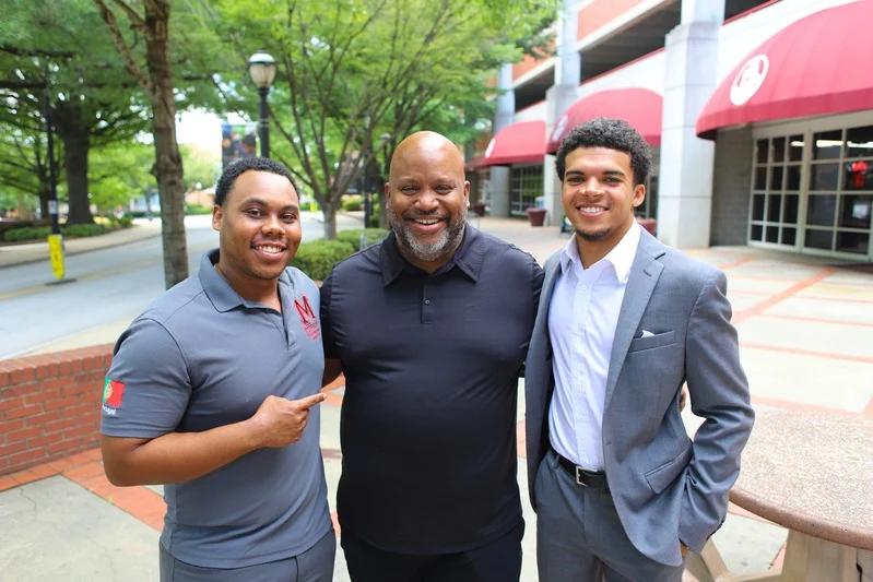 Three men smiling in an outdoor campus setting.
