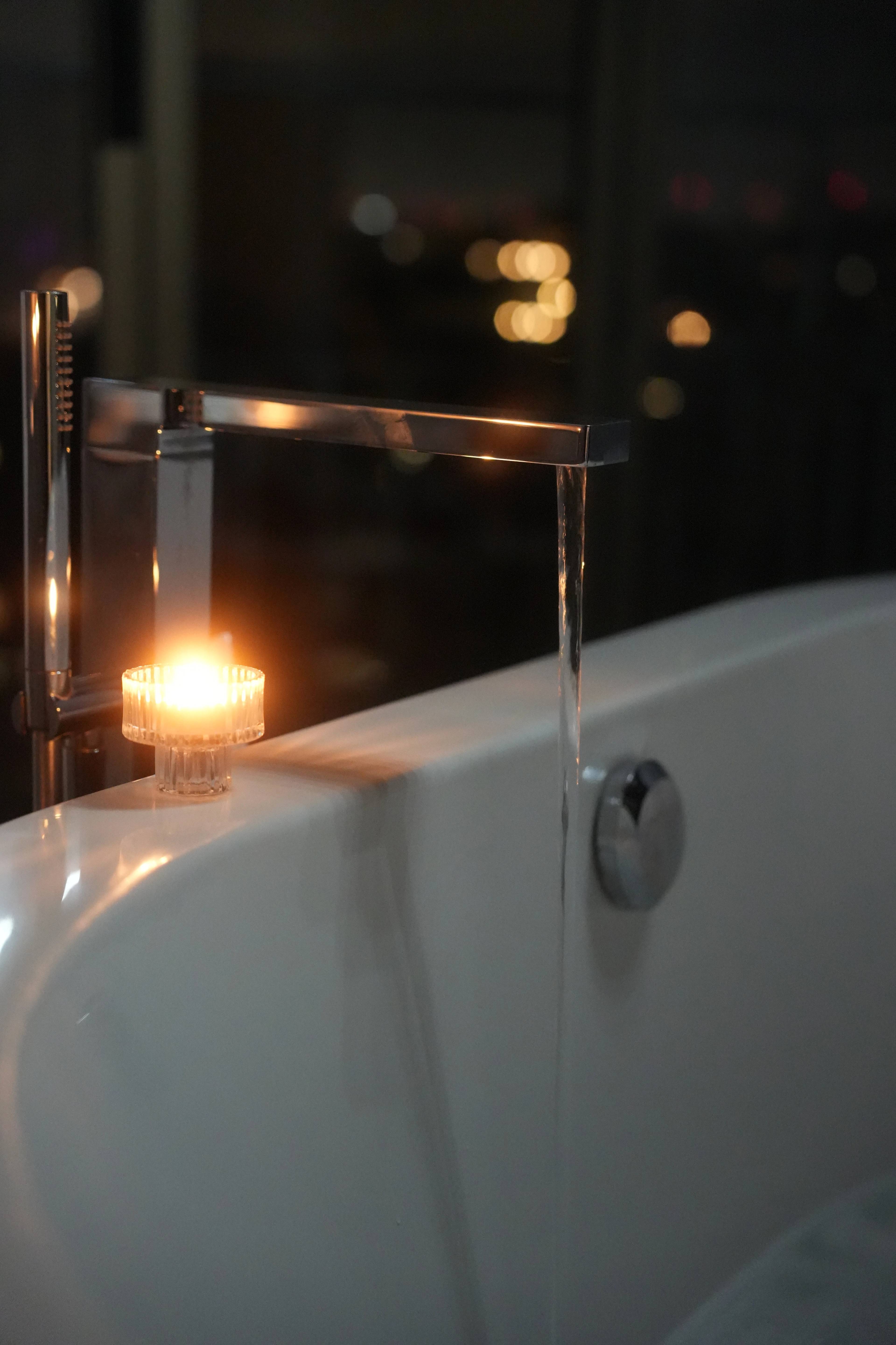 Brass tap pouring water into the tub, a single candle on the rim, city bokeh outside