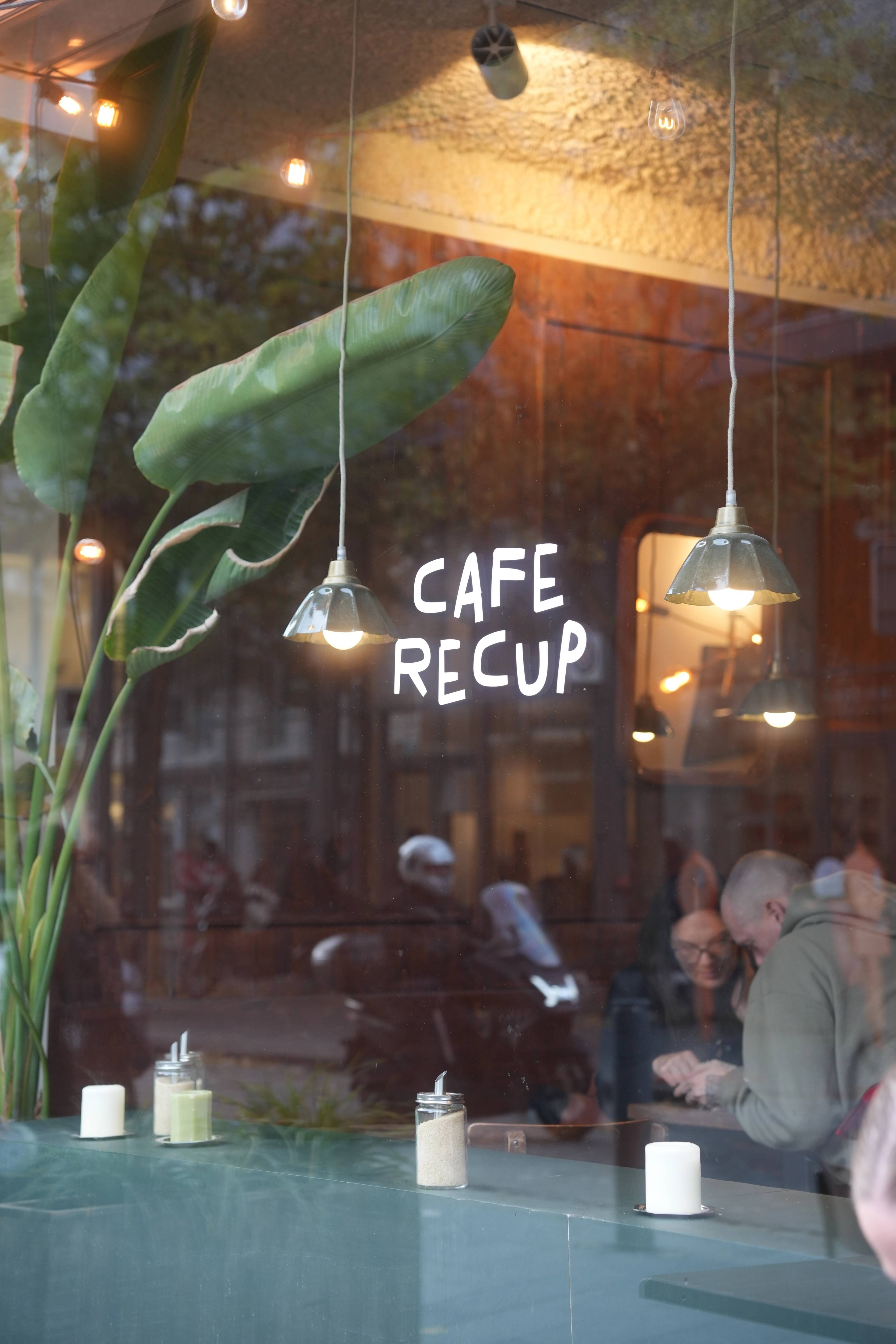 Café Recup window — pendant lights, banana plants, a couple at the counter inside