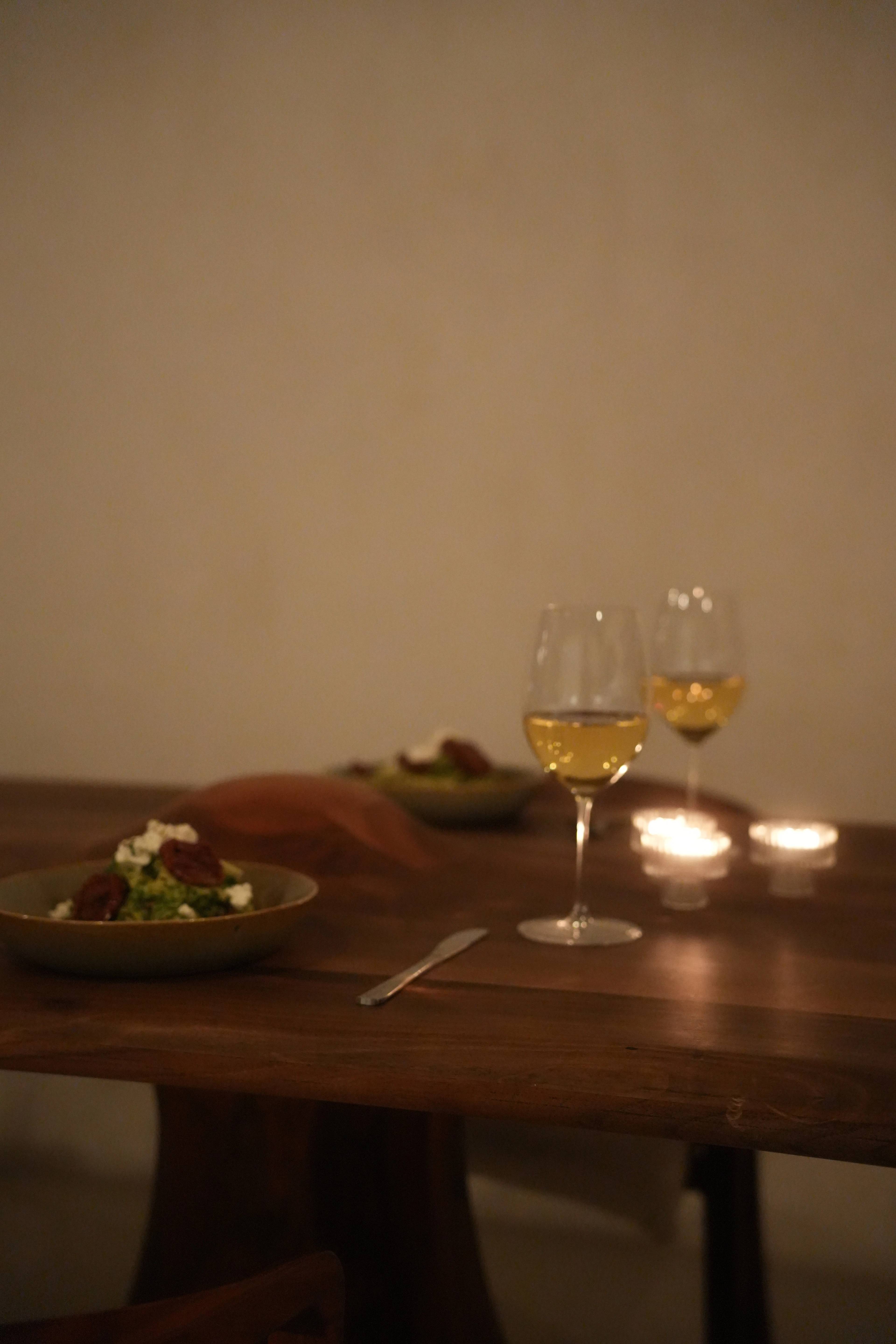 Two plates of greens with feta and dates, two glasses of white wine, candles between them