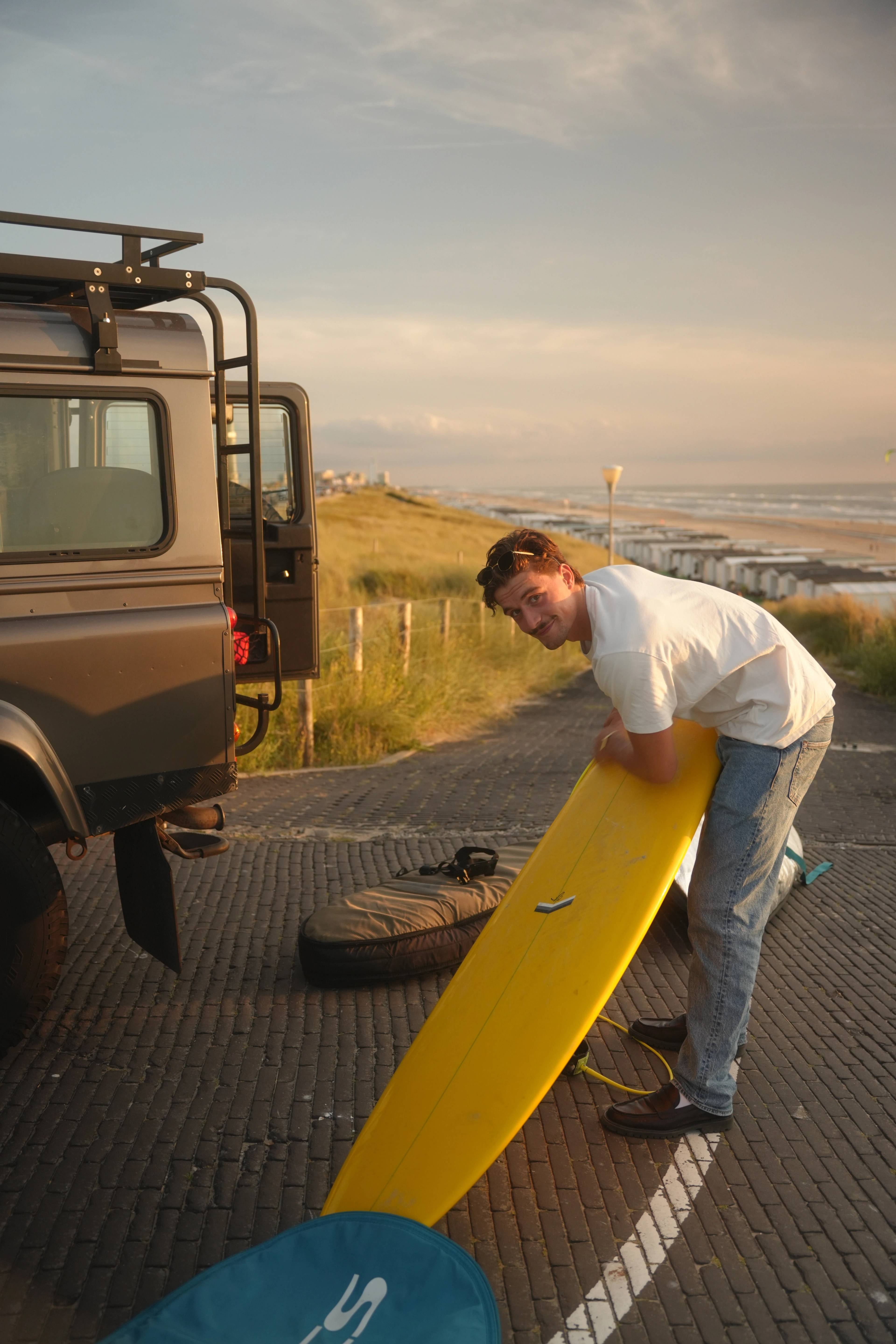 Waxing a yellow surfboard at golden hour next to the Landrover, dunes behind