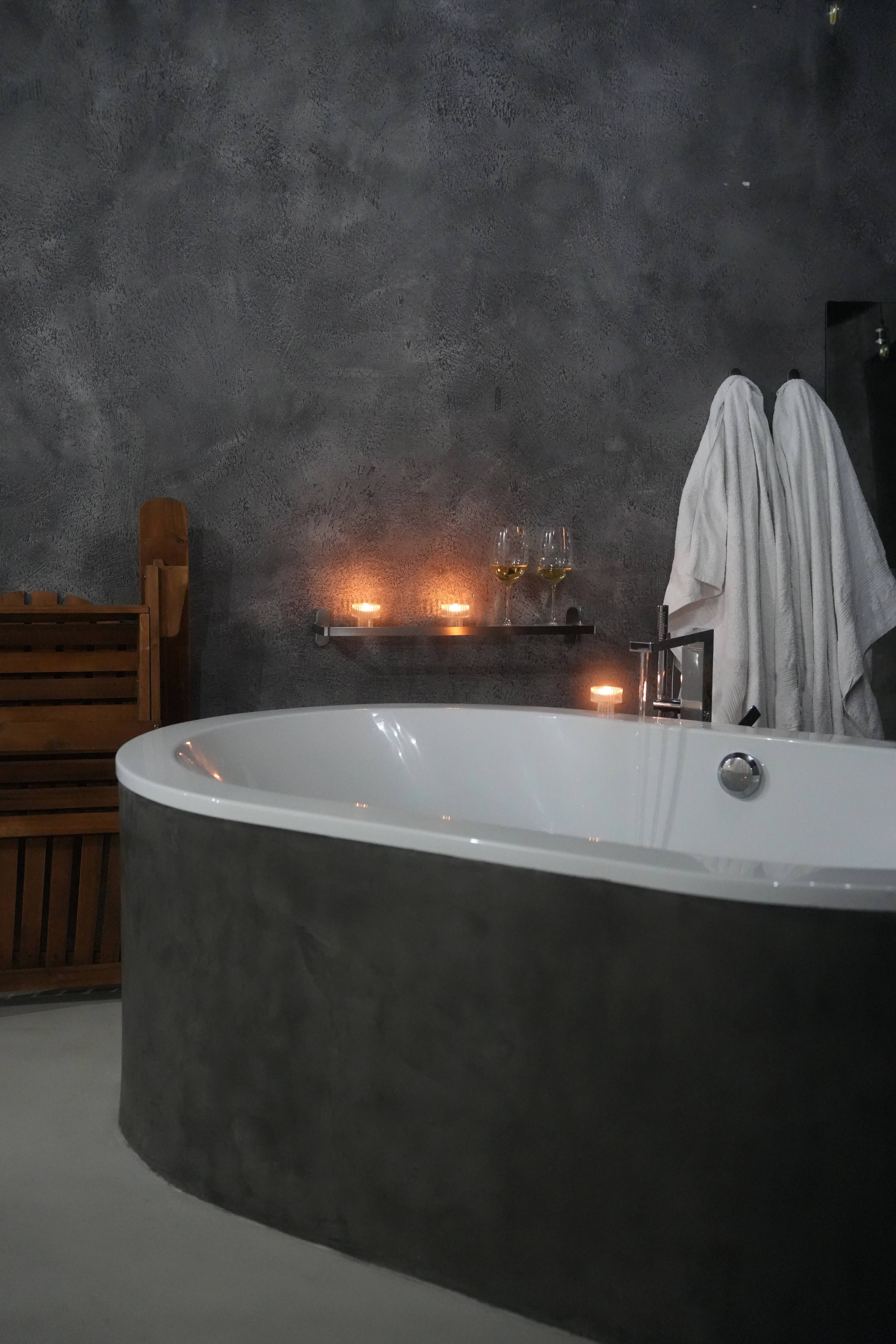 Wide tub against a concrete wall, candles on a shelf, two white robes hung beside