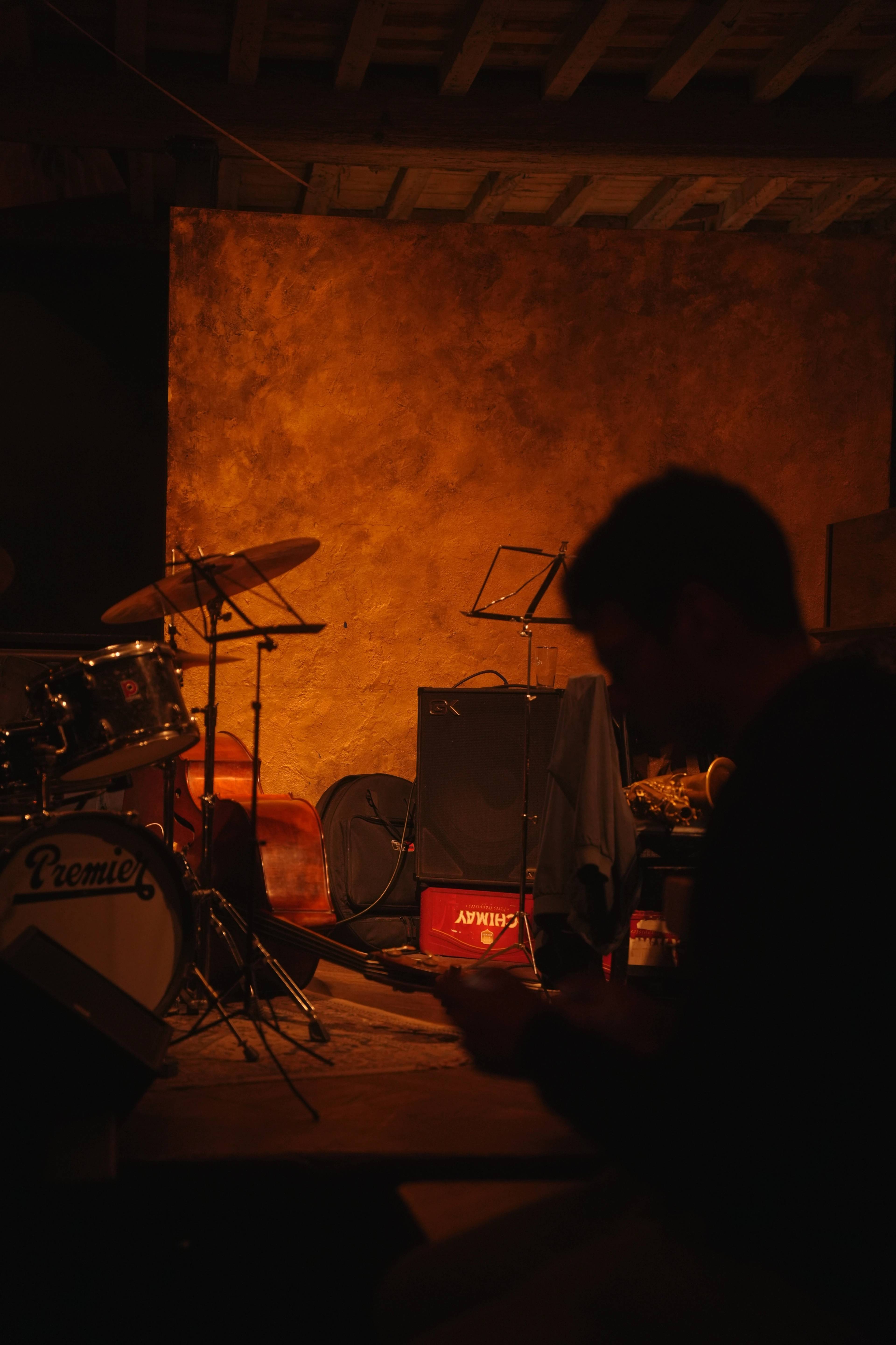 Dim red-lit corner of a small venue — drum kit, Chimay crate, someone reading in silhouette