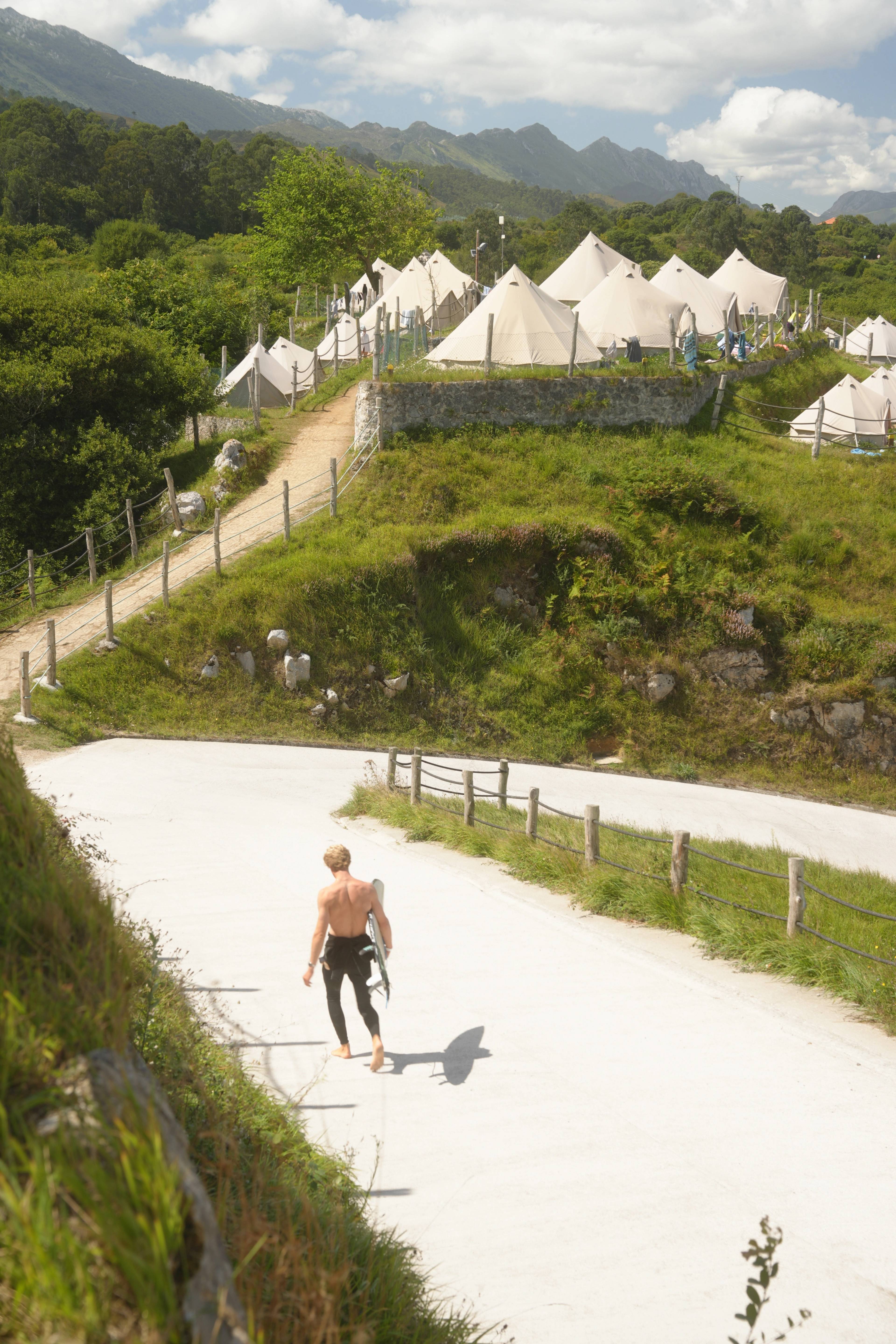 Bell tents arranged in the green valley below the mountains, a surfer walking back to camp
