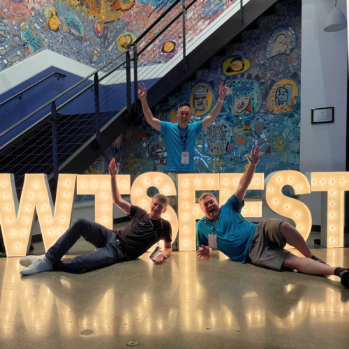 Three members of the Nozzle team pose in front of illuminated WTSFest letters and a colourful mosaic wall. Boyd (left) and Derek (right) recline playfully on the floor with their arms raised, while Duncan stands behind the letters with both hands raised in peace signs.
