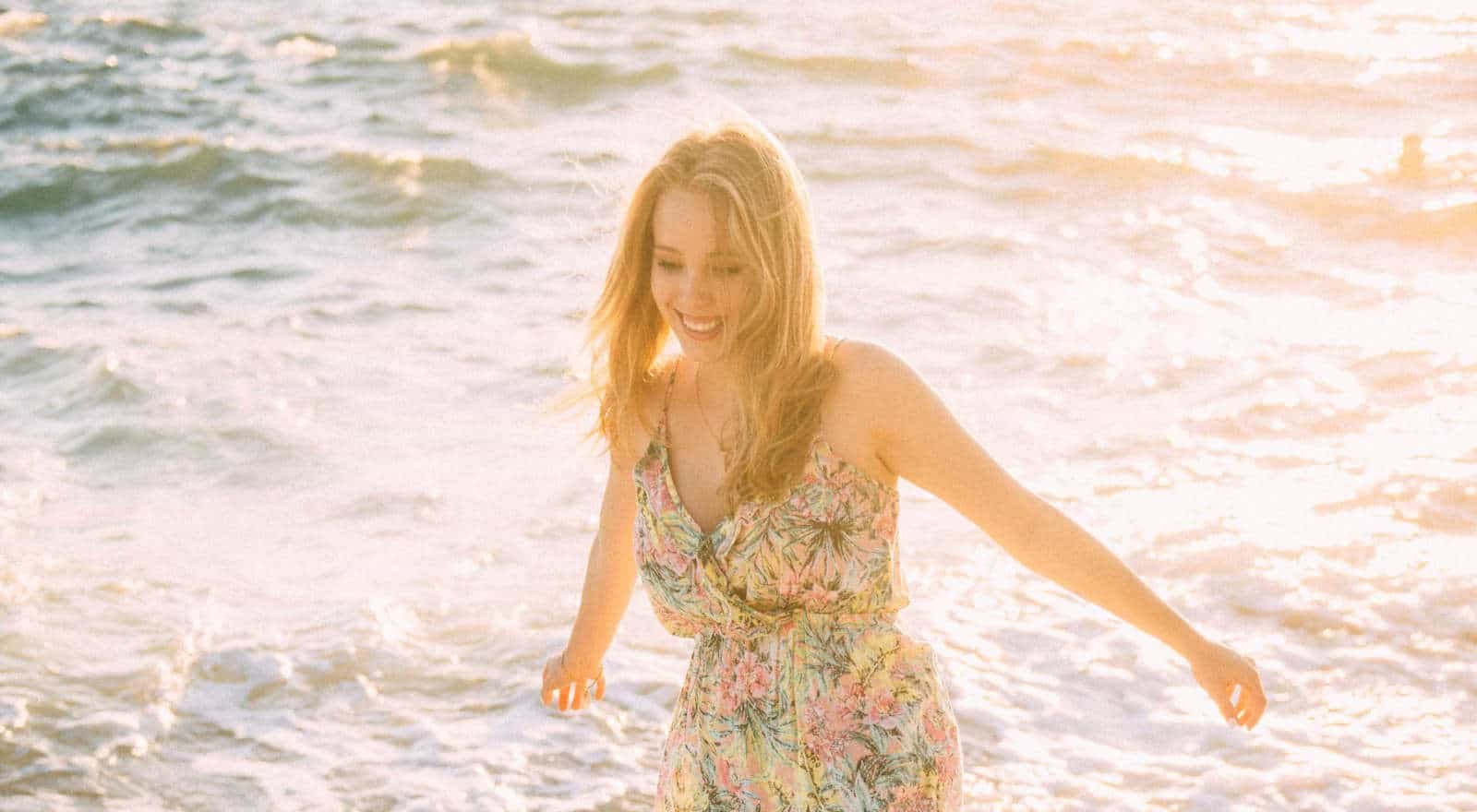 A smiling woman with long fair hair in a floral sundress wading in the sea in bright evening sunlight