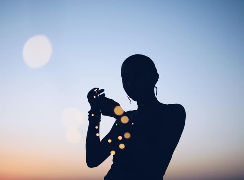 Silhouette of a woman holding a string of small glowing lights against a dusk sky