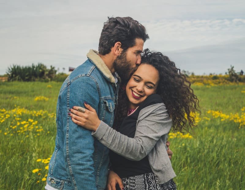 A bearded man in a denim jacket kissing the head of a laughing woman with curly hair as they hug in a meadow of yellow wildflowers