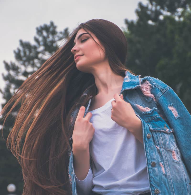 A young woman with her eyes closed and long brown hair blowing in the wind, wearing a white top and a ripped denim jacket, with pine trees behind her