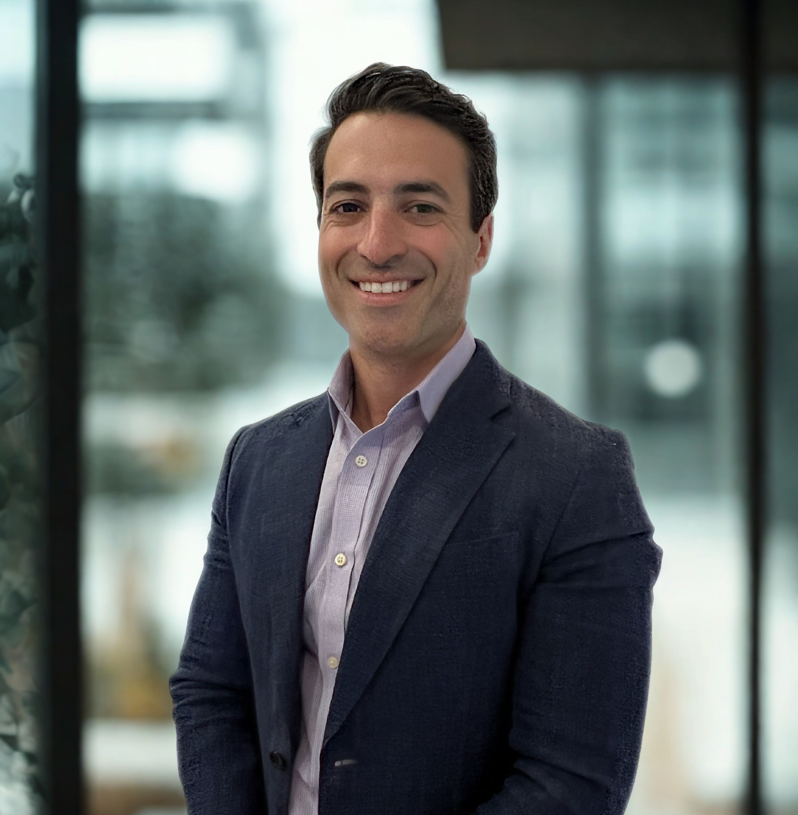 Professional headshot of Craig Langan, a man with short dark hair smiling warmly, wearing a navy blazer over a light blue button-down shirt against a blurred office background.