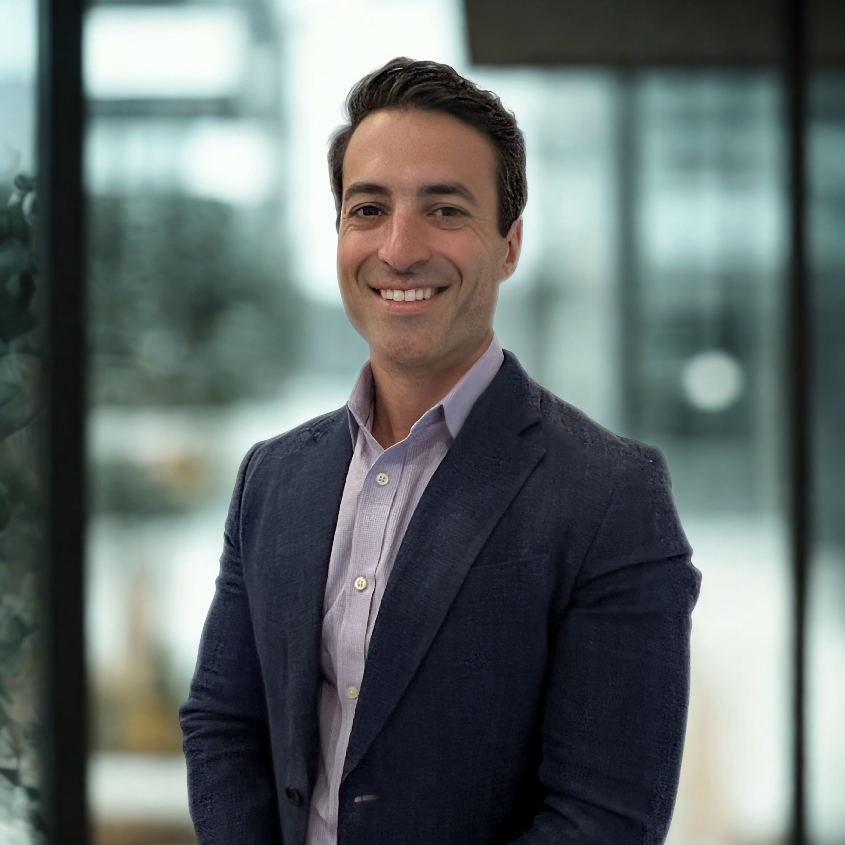 Professional headshot of Craig Langan, a man with short dark hair smiling warmly, wearing a navy blazer over a light blue button-down shirt against a blurred office background.