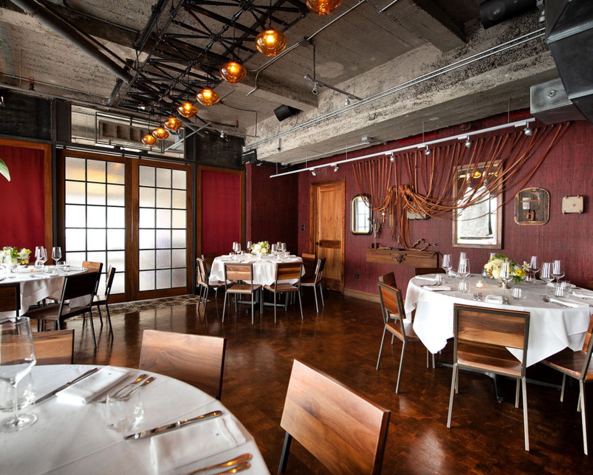 View of the 57 Room, with multiple round tables, exposed concrete ceiling, wood floors, and rustic decor on dark red walls