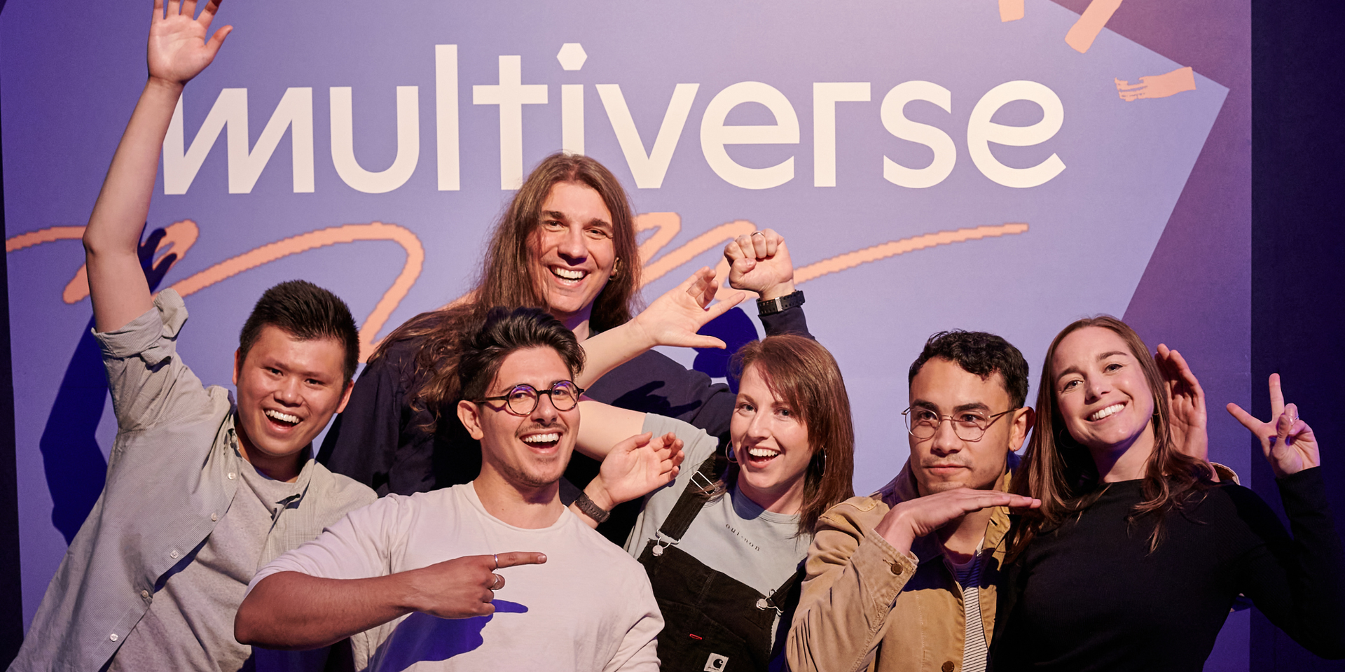 A group of employees striking a funny pose smiling, in front of a Multiverse banner