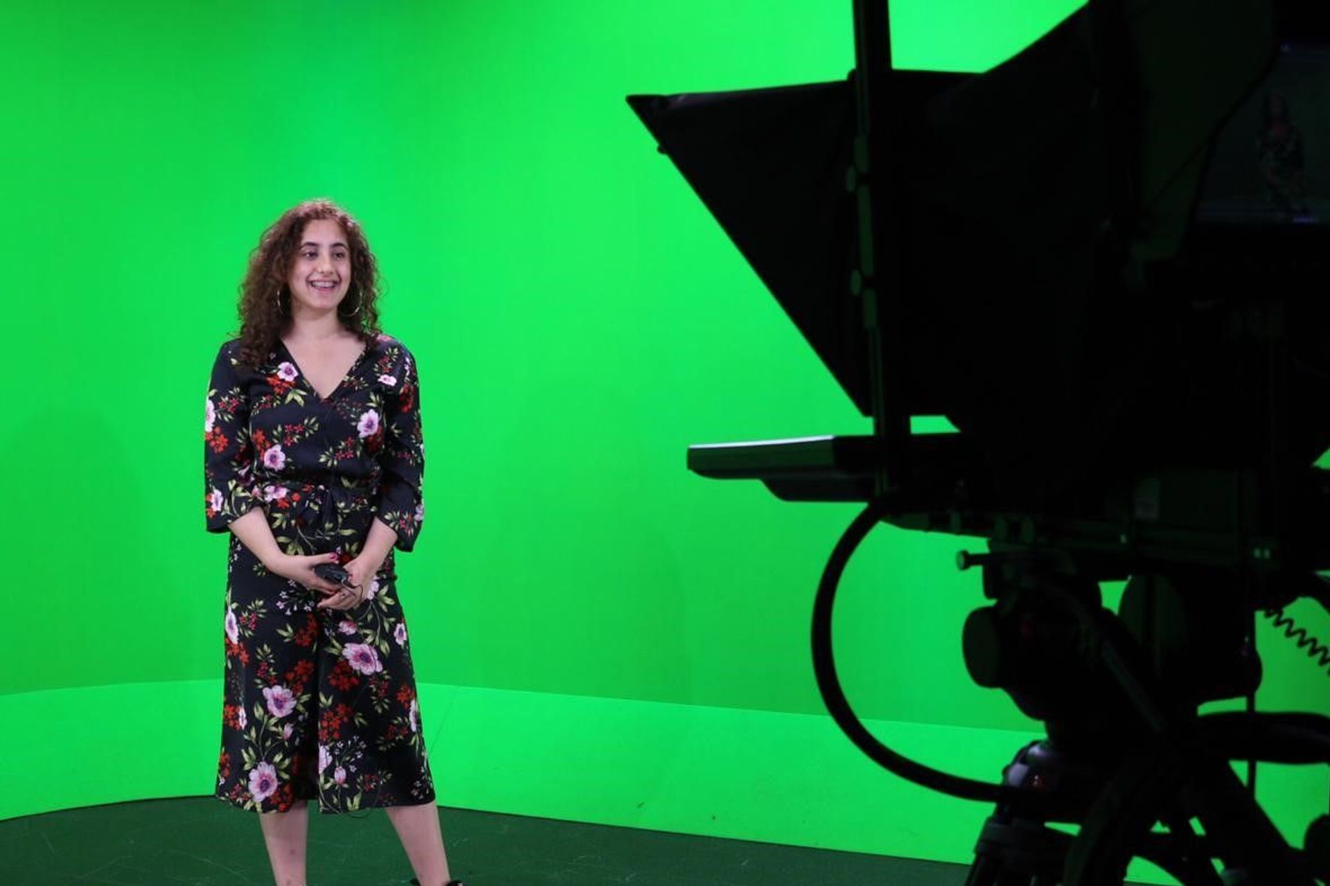 An image of a woman in a flowery dress standing in front of a film camera smiling in a green screen room