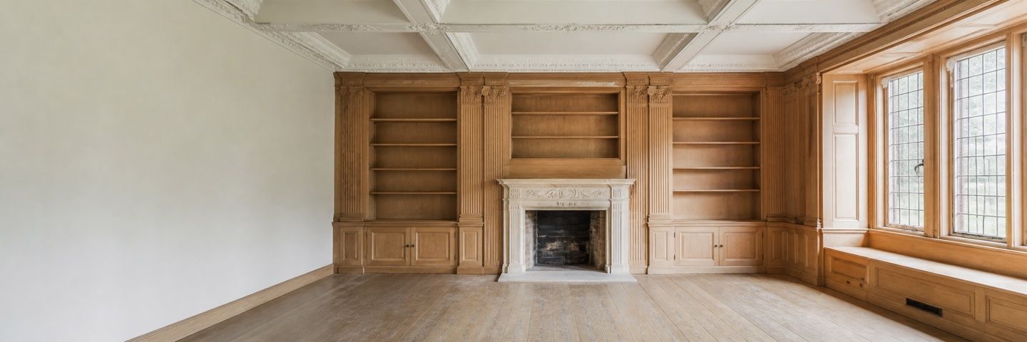 Illustration — An empty panelled library. Oak bookcases with fluted pilasters stand either side of a carved white stone fireplace, under a coffered plaster ceiling with a moulded cornice. Leaded casement windows and a built-in window seat fill the right-hand wall; the left wall is bare.