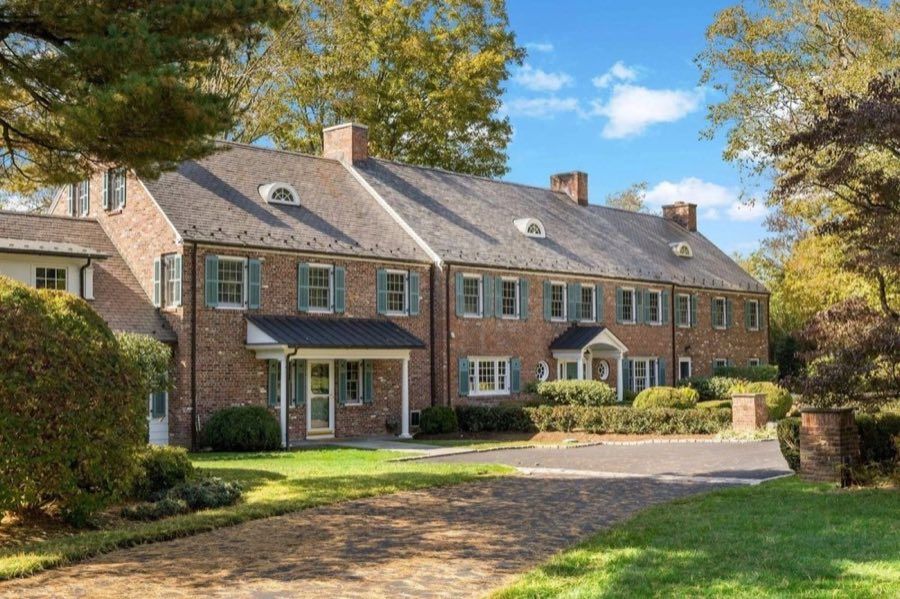 Long brick colonial with a slate gambrel roof, arched dormer windows, pale green shutters and two entry porticoes, beside a cobbled forecourt.