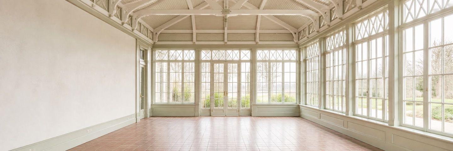 Illustration — An empty conservatory glazed on three sides with tall lattice-headed windows and french doors, looking out to lawn and bare winter trees. A painted timber roof truss spans overhead; the floor is terracotta tile. The left-hand wall is plain plaster.