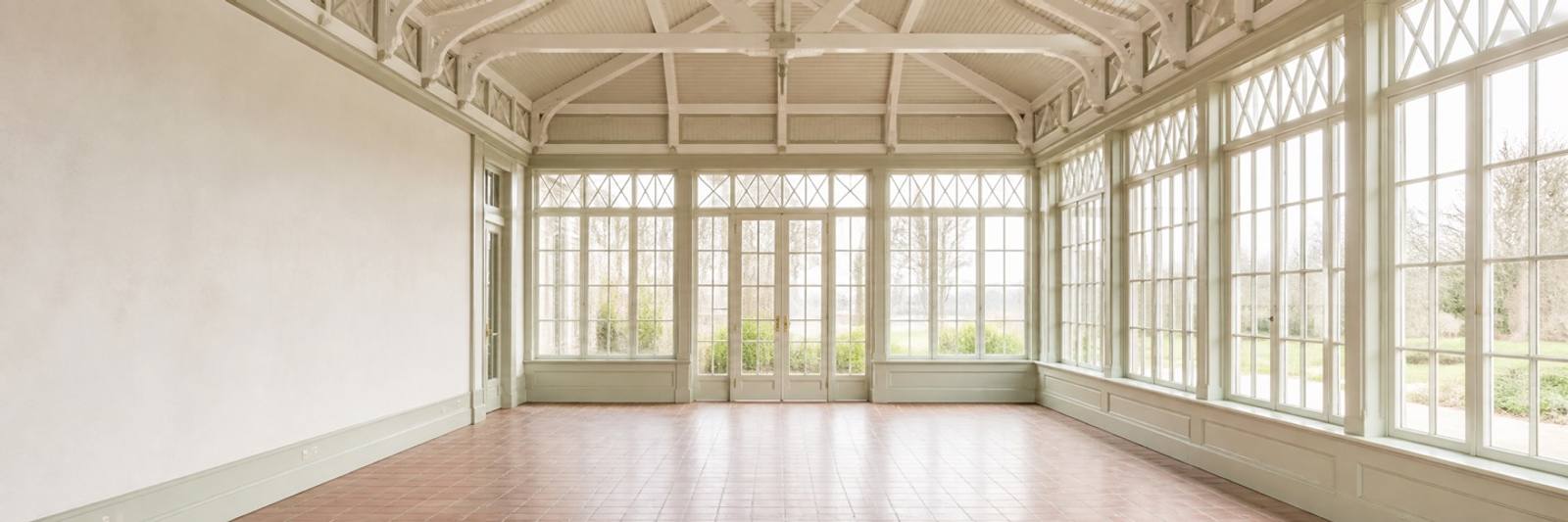 Illustration — An empty conservatory glazed on three sides with tall lattice-headed windows and french doors, looking out to lawn and bare winter trees. A painted timber roof truss spans overhead; the floor is terracotta tile. The left-hand wall is plain plaster.