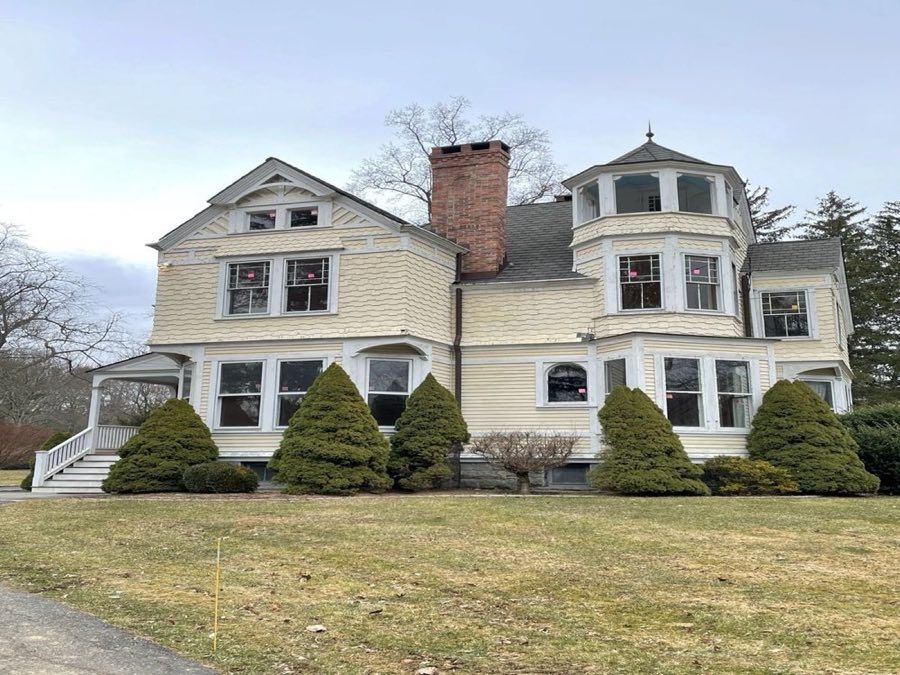 Pale yellow Queen Anne Victorian with a polygonal corner tower, a decorated gable, brick chimney and scalloped shingles, with conical clipped shrubs along the front in winter.