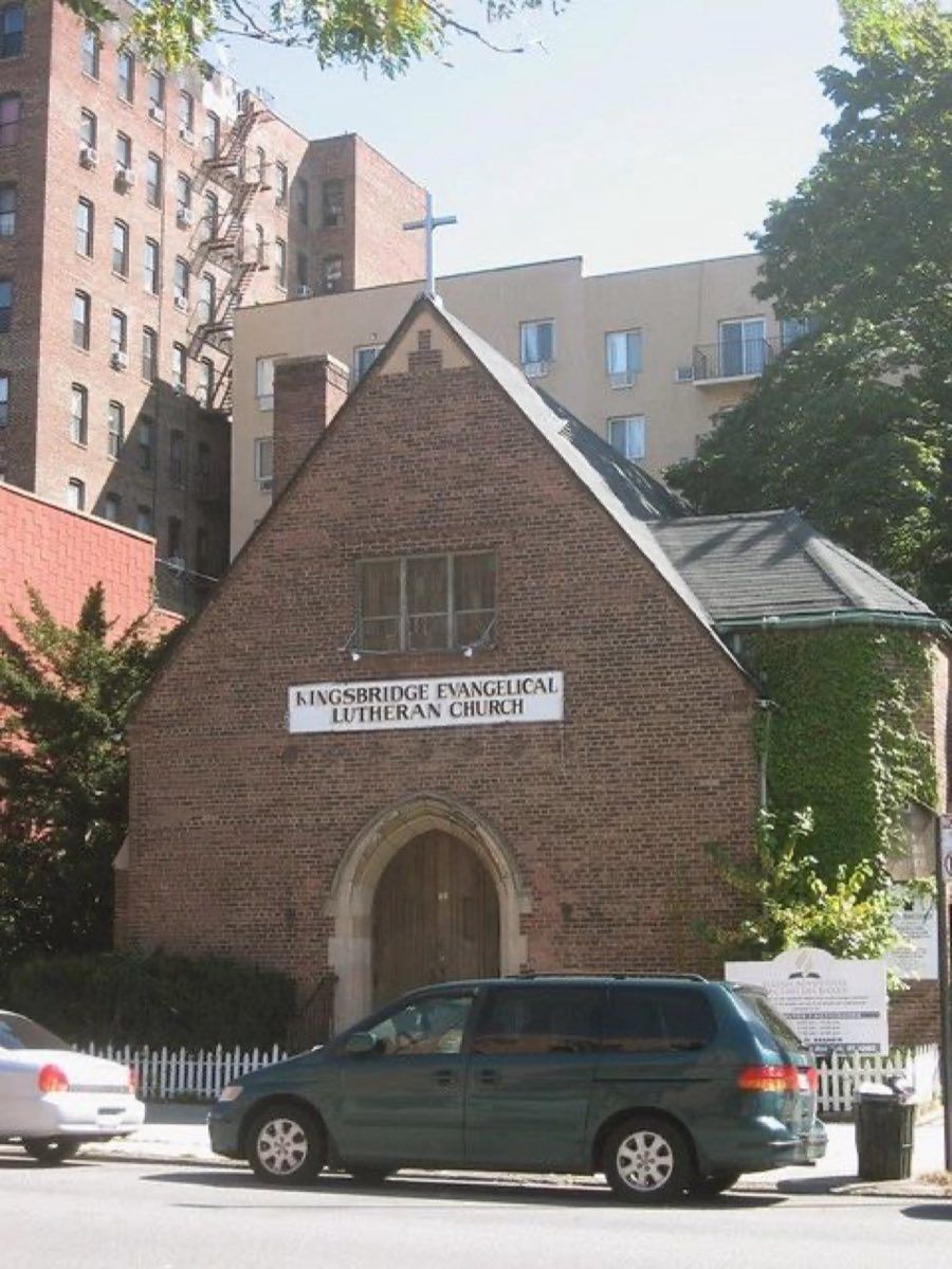 Brick church with a steep gabled front, an arched wooden door and a rooftop cross, signed Kingsbridge Evangelical Lutheran Church, between apartment buildings on a city street.