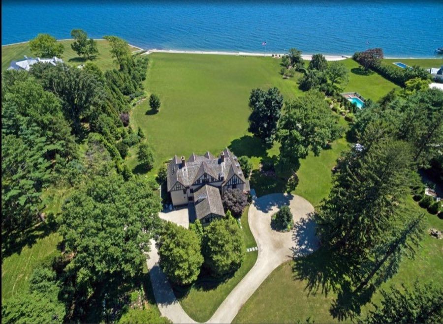 Aerial view of a half-timbered Tudor-style house among mature trees, with a wide lawn running down to a private shoreline and a swimming pool beyond.