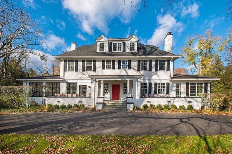 White stucco colonial with black shutters, a columned entry porch with a red door and glassed sun porches to either side, under bare autumn branches.