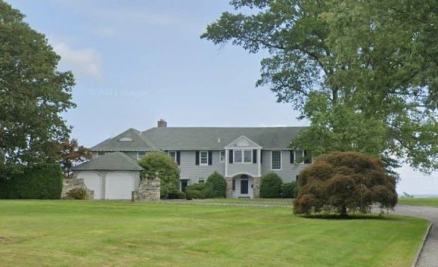 Grey shingled house with a pedimented entry, attached garage and stone base, on a wide lawn with a copper beech, water visible beyond.