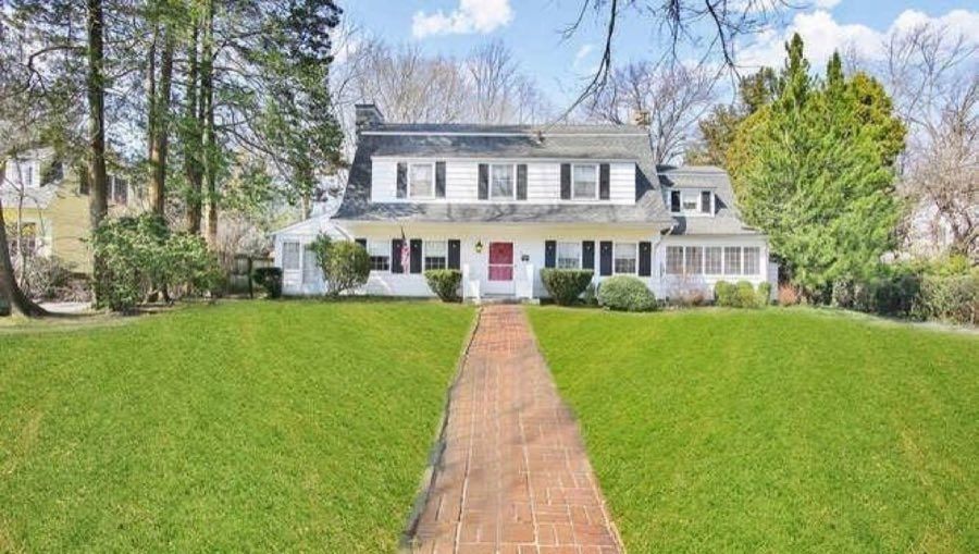 White Dutch colonial with a gambrel roof, black shutters and a red front door, at the end of a straight brick path across a level lawn.