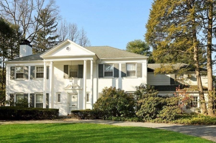 White painted brick colonial with a two-storey columned portico, grey shutters and a pedimented door surround, behind clipped hedging and lawn.