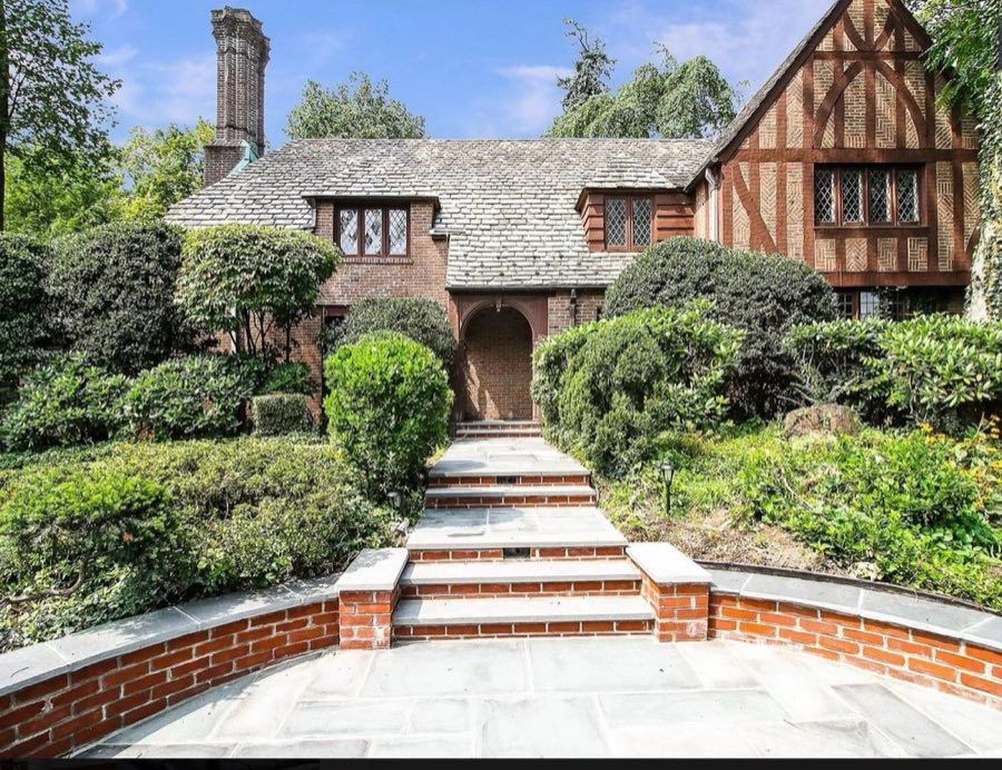 Brick and half-timbered Tudor with a steep slate roof, a tall patterned chimney, leaded windows and an arched entry, above brick-edged bluestone steps.