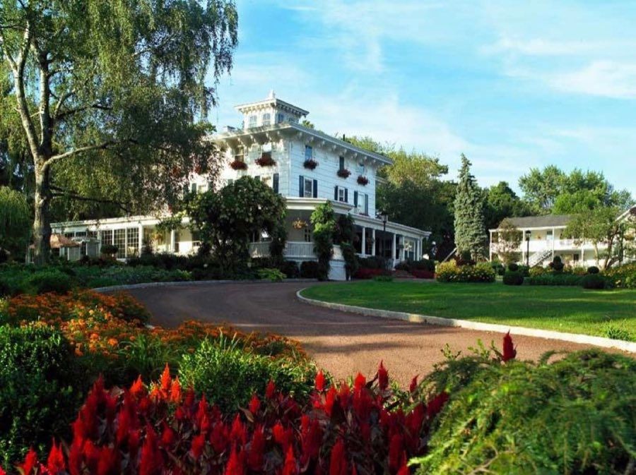 Large white Italianate inn with a rooftop cupola, deep verandas, window boxes and shutters, at the head of a curving drive behind beds of red and orange flowers.