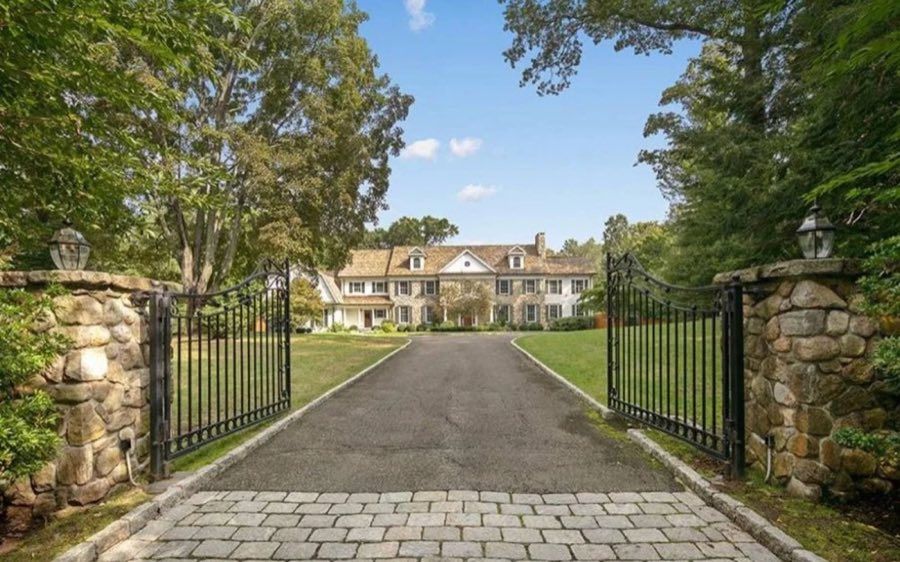 View through open wrought-iron gates set in fieldstone piers, up a cobble-edged drive to a large shingled colonial with dormer windows.